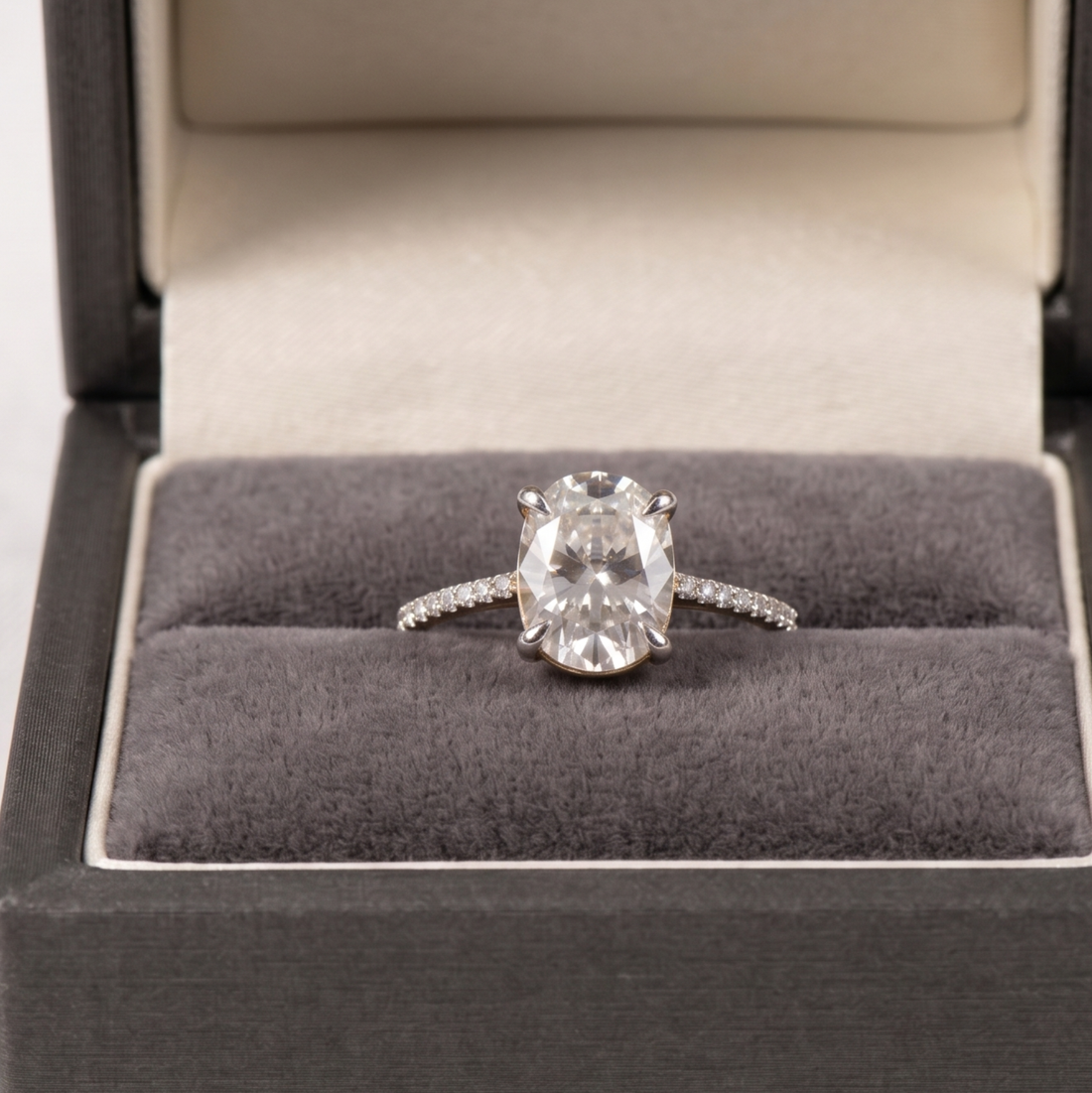 An oval-cut diamond ring in a silver-toned solitaire setting, displayed inside an open dark gray velvet ring box.