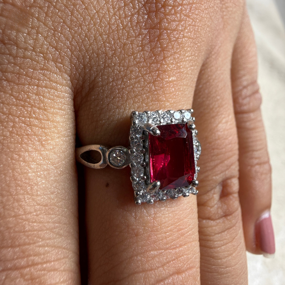 A silver-toned ring with a rectangular red gemstone surrounded by small clear stones, worn on a finger.