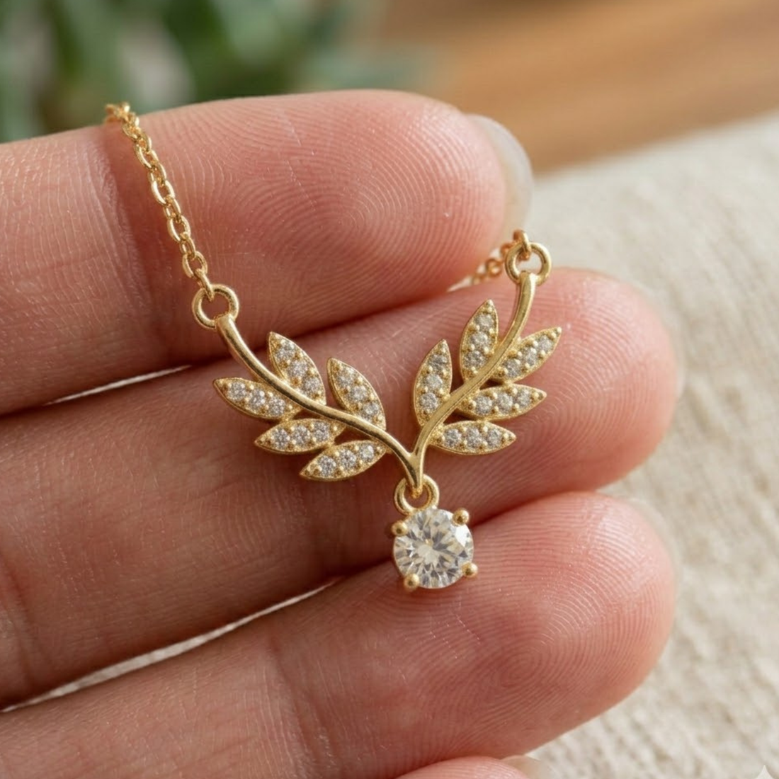 A gold leaf pendant necklace with clear stones and a round clear gemstone, held on a person's fingers against a soft beige background.