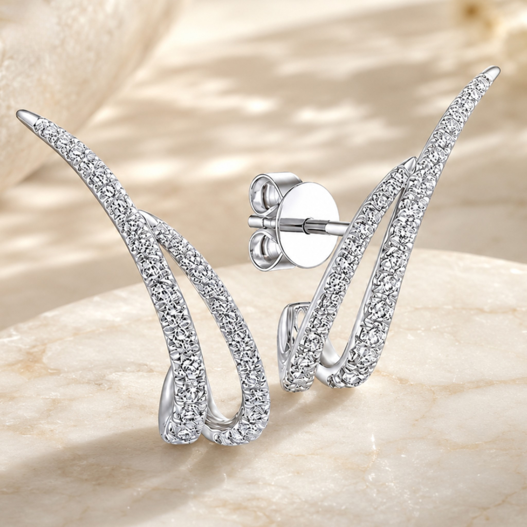A pair of silver-toned wing-shaped stud earrings paved with clear cubic zirconia stones, displayed on a light beige stone surface. One earring is shown from the front, and the other is angled to show the post and butterfly back.