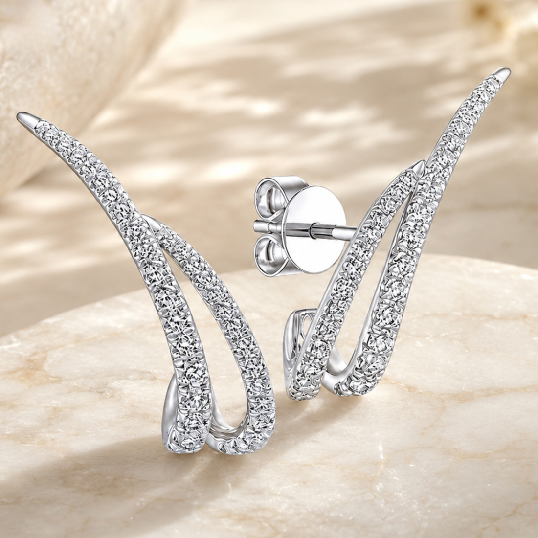 A pair of silver-toned wing-shaped stud earrings paved with clear cubic zirconia stones, displayed on a light beige stone surface. One earring is shown from the front, and the other is angled to show the post and butterfly back.