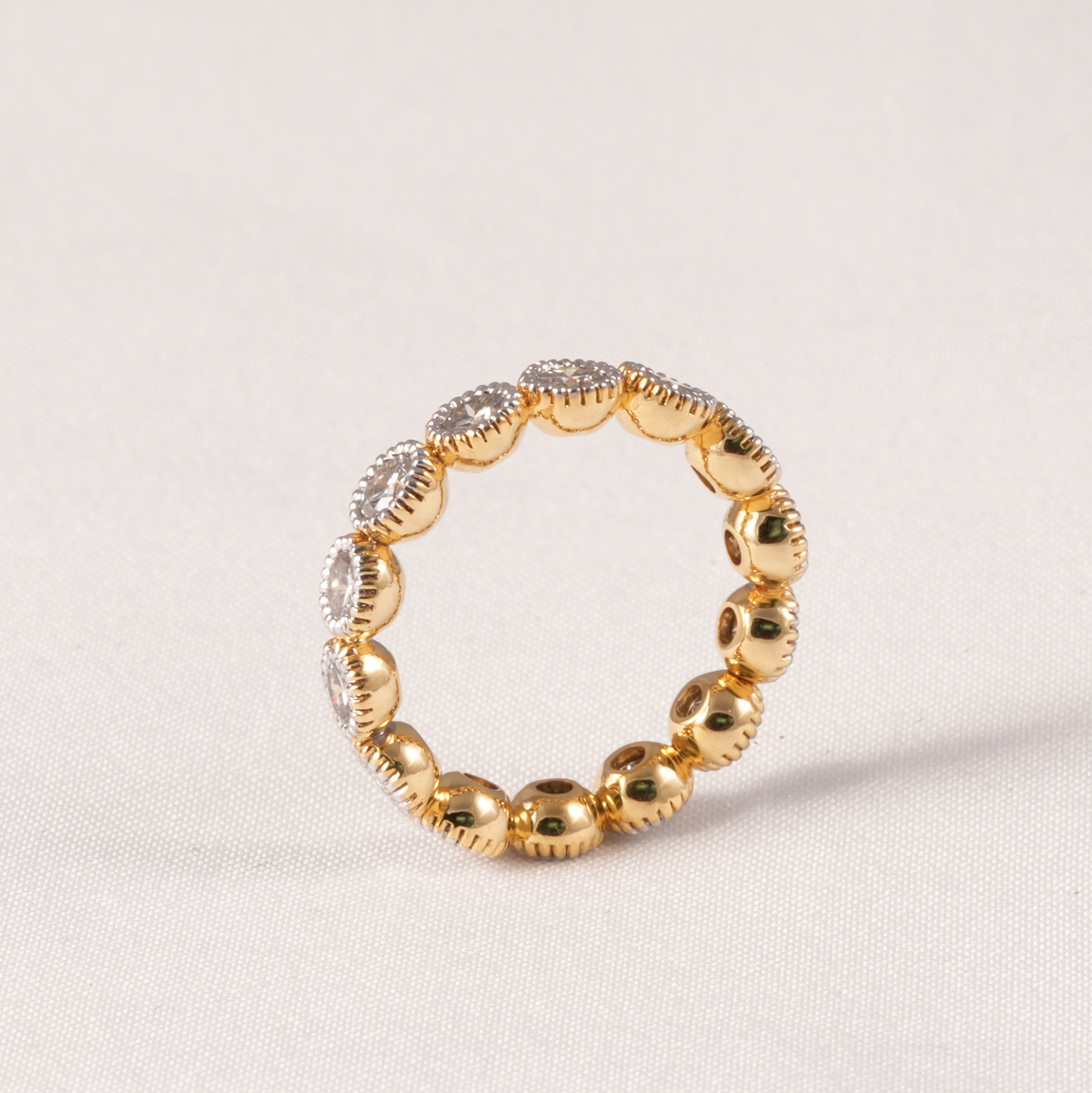 A gold beaded ring with alternating polished round beads and textured oval beads set with clear stones, photographed on a light beige fabric background.