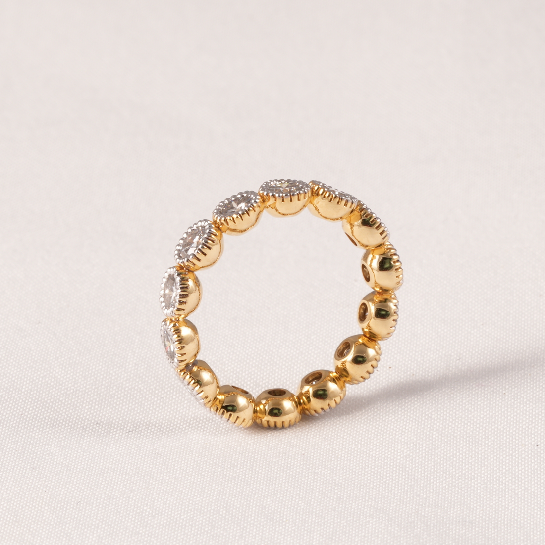 A gold beaded ring with alternating polished round beads and textured oval beads set with clear stones, photographed on a light beige fabric background.
