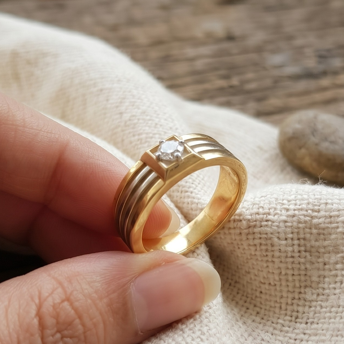 A gold ring with a single clear round gemstone held between fingers over a textured beige fabric background.