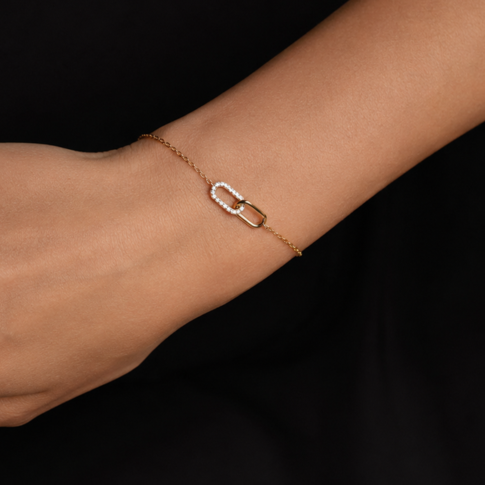 A delicate gold chain bracelet with a small interlocking open-link design, one side of which is lined with clear stones, shown worn on a person's wrist against a black background.