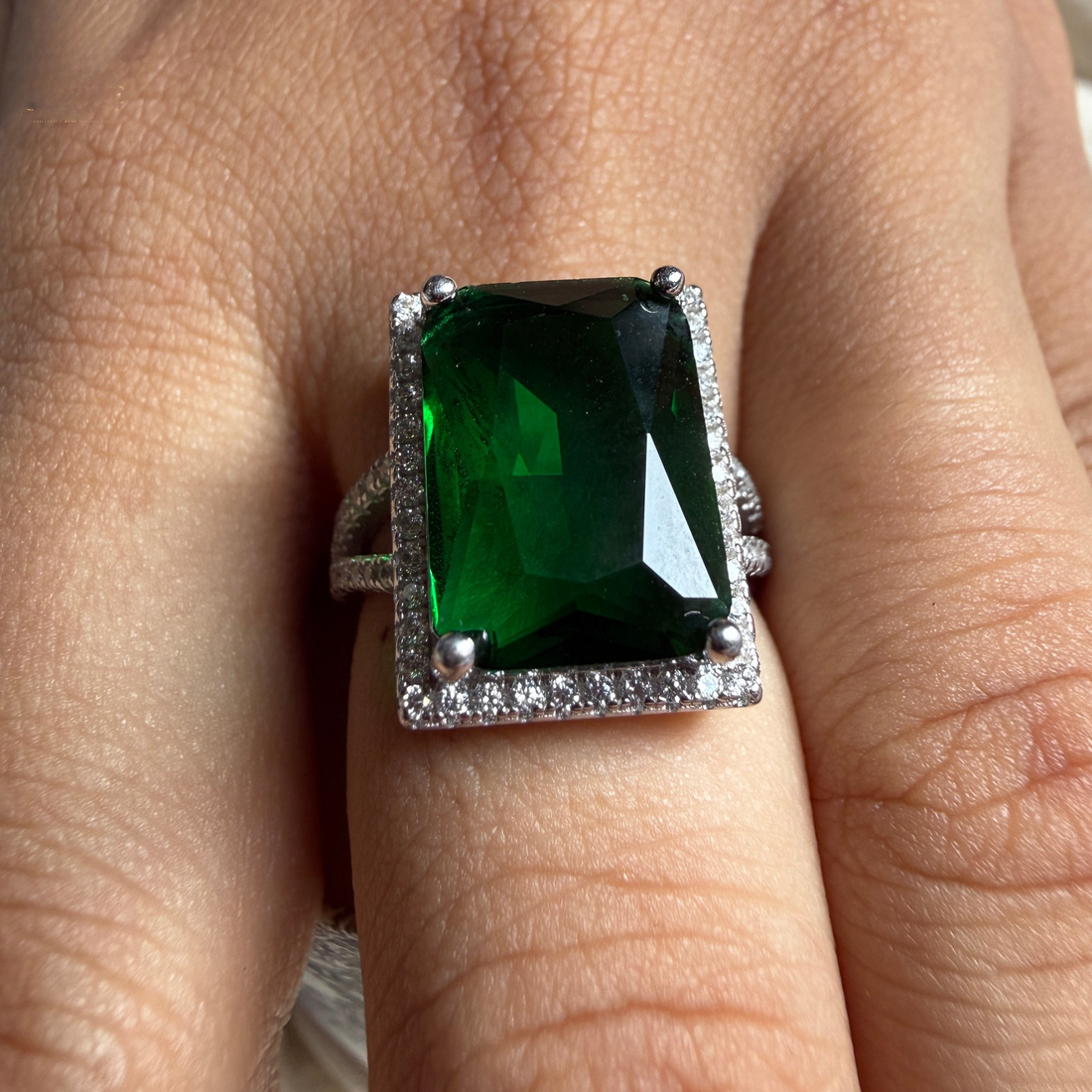 A rectangular emerald-cut green gemstone ring with a silver-toned halo and small clear accent stones, shown worn on a finger.