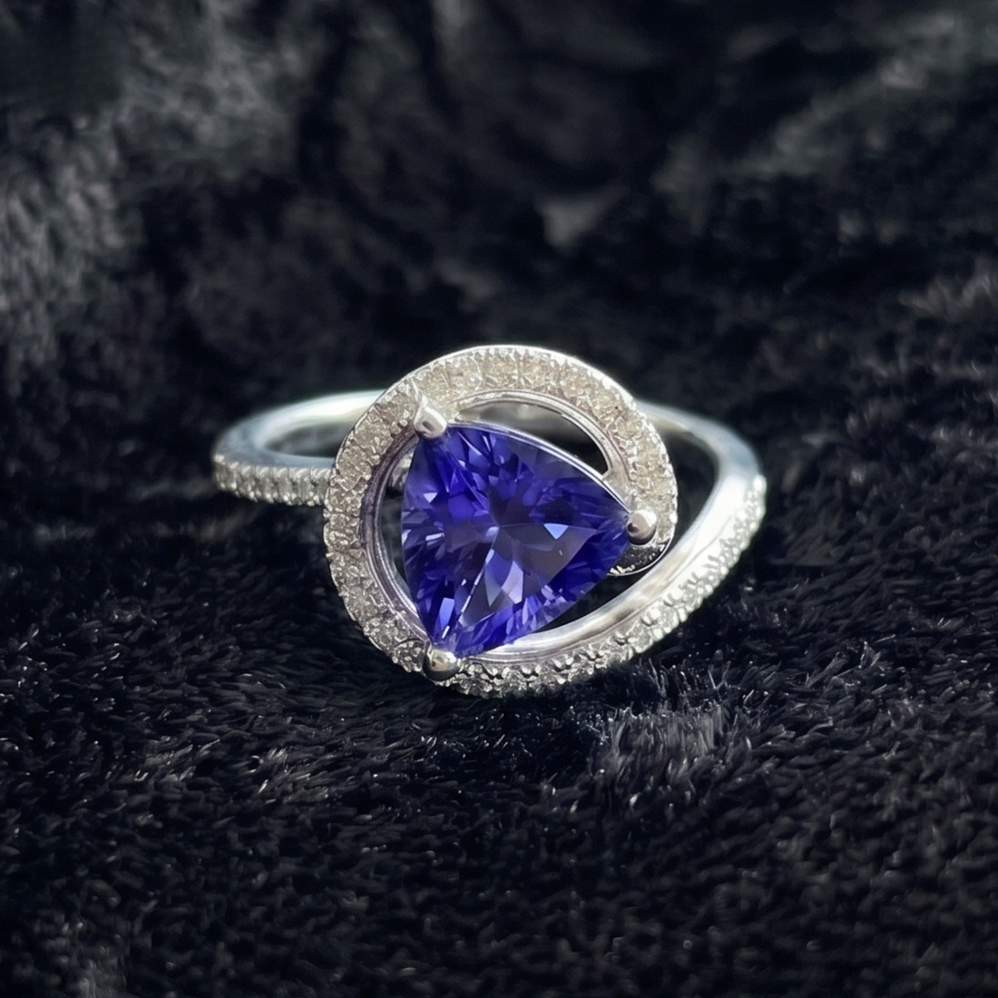 A silver ring featuring a pear-cut purple tanzanite center stone surrounded by a double halo of small clear stones, displayed on a textured black fabric background.