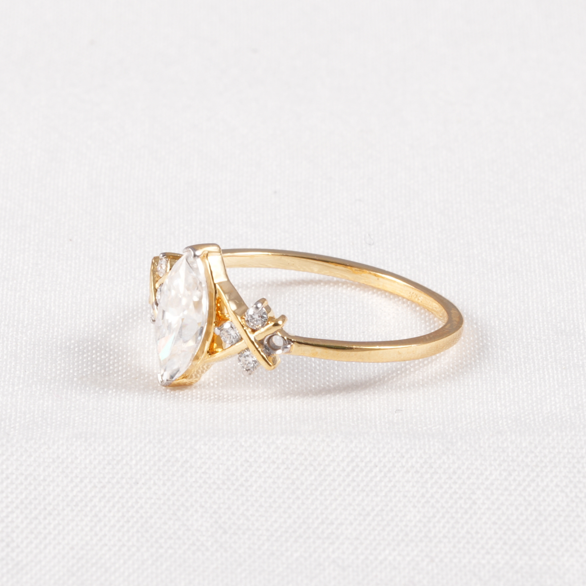 A gold ring featuring a marquise-cut clear gemstone and small accent diamonds, set on a textured white background.