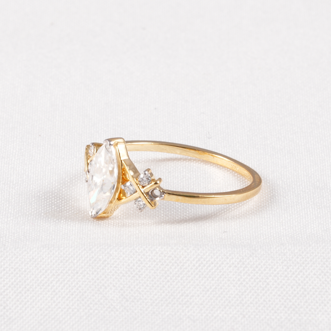 A gold ring featuring a marquise-cut clear gemstone and small accent diamonds, set on a textured white background.