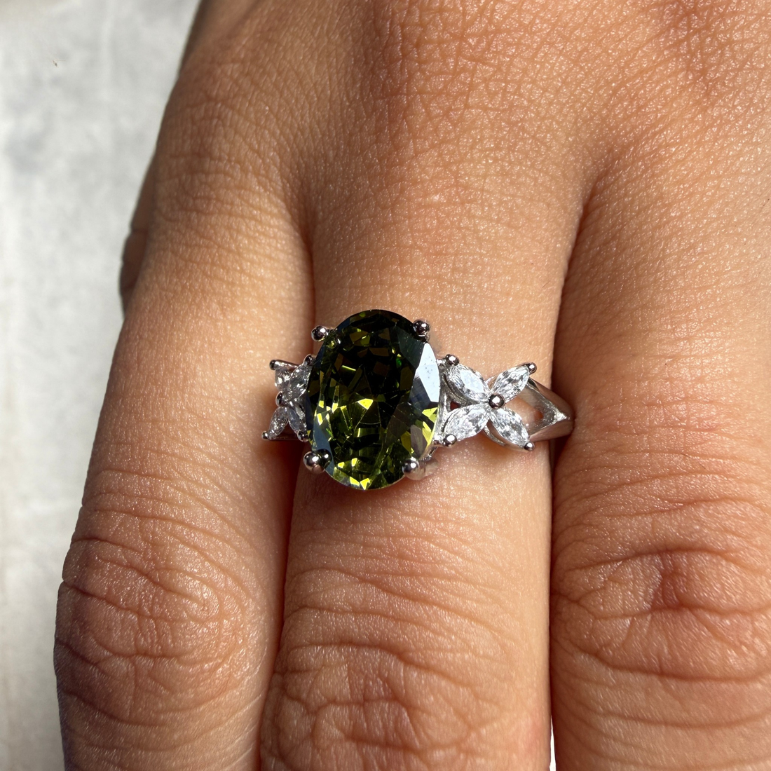A silver ring with a large oval green gemstone and small clear accent stones, shown worn on a finger.