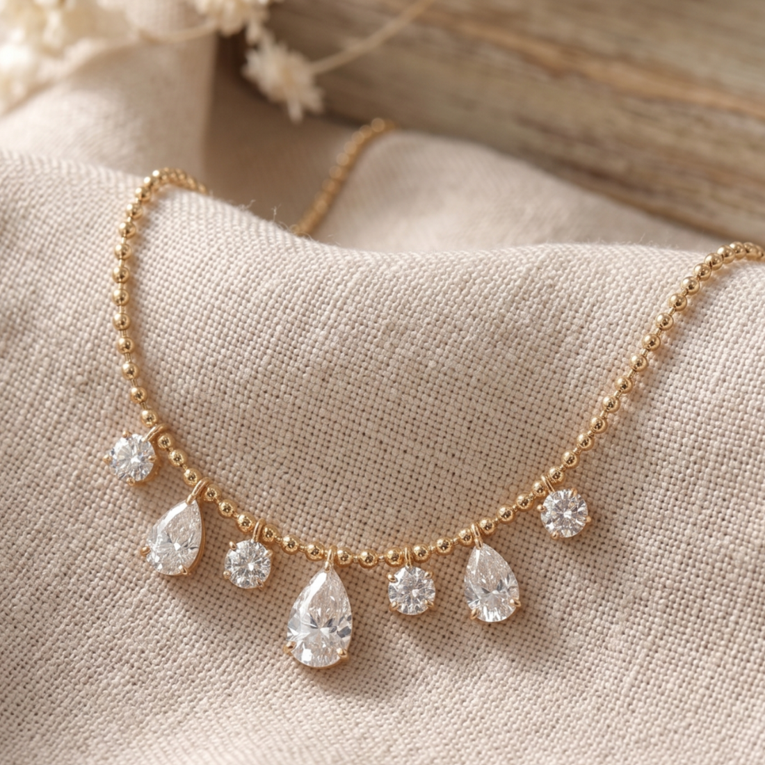 A delicate gold chain necklace with five dangling clear teardrop and round gemstones, displayed on a textured beige fabric surface.