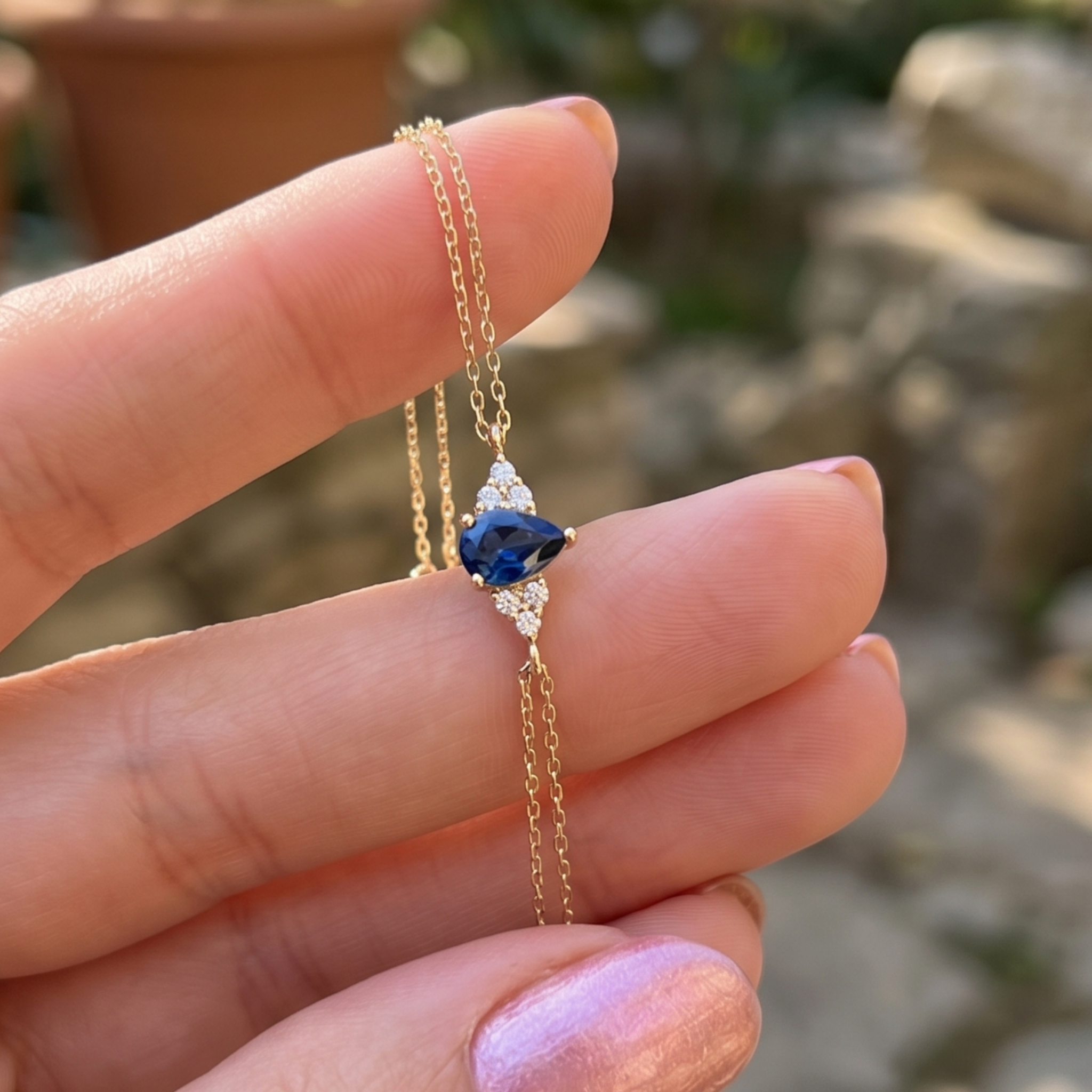 A delicate gold chain necklace with a central blue teardrop-shaped gemstone and small clear accent stones, held between fingers against a blurred outdoor background.