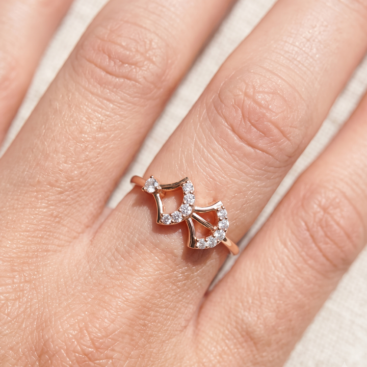 A close-up of a hand wearing a rose gold ring with a double-heart design set with small clear stones.