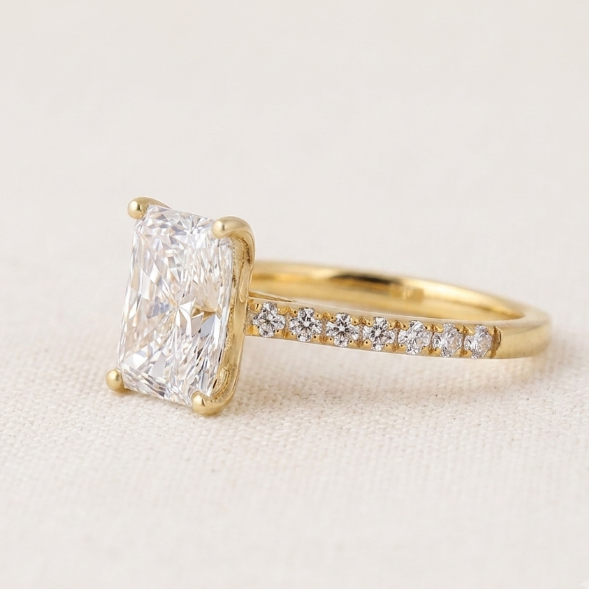 A yellow gold ring featuring a large rectangular radiant-cut diamond solitaire and a pavé band with small round diamonds, set against a light beige textured background.
