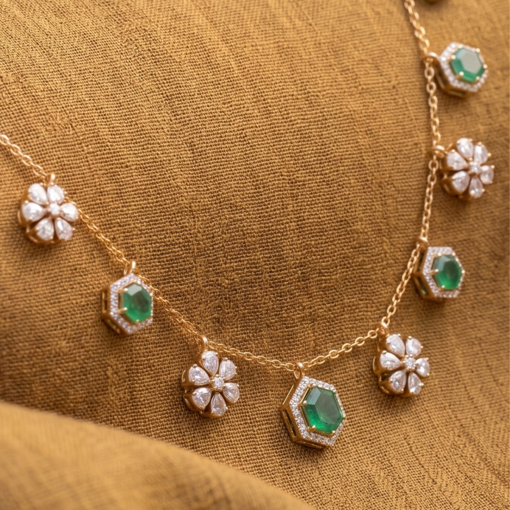 A gold chain necklace featuring alternating green emerald-cut stones and white flower-shaped pavé settings, displayed on a textured gold fabric background.