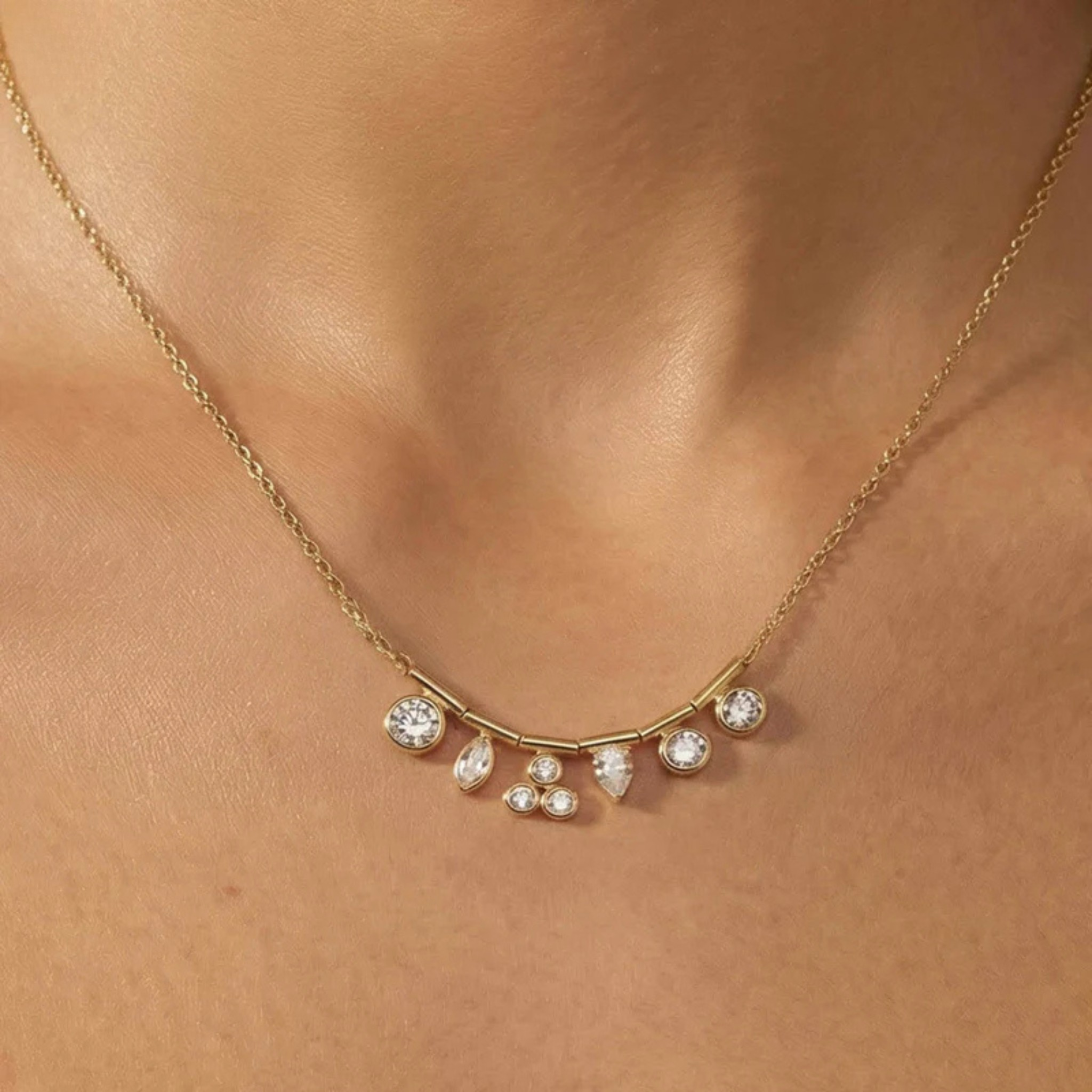 A delicate gold necklace with a curved bar and small clear teardrop and round stones, shown worn on a person's neck.