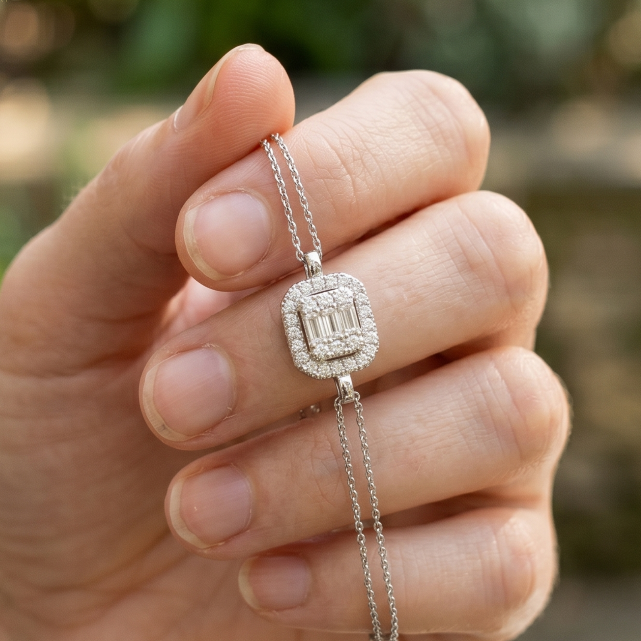 A close-up of a hand holding a silver-toned pendant necklace with a rectangular baguette-cut center stone surrounded by smaller stones.