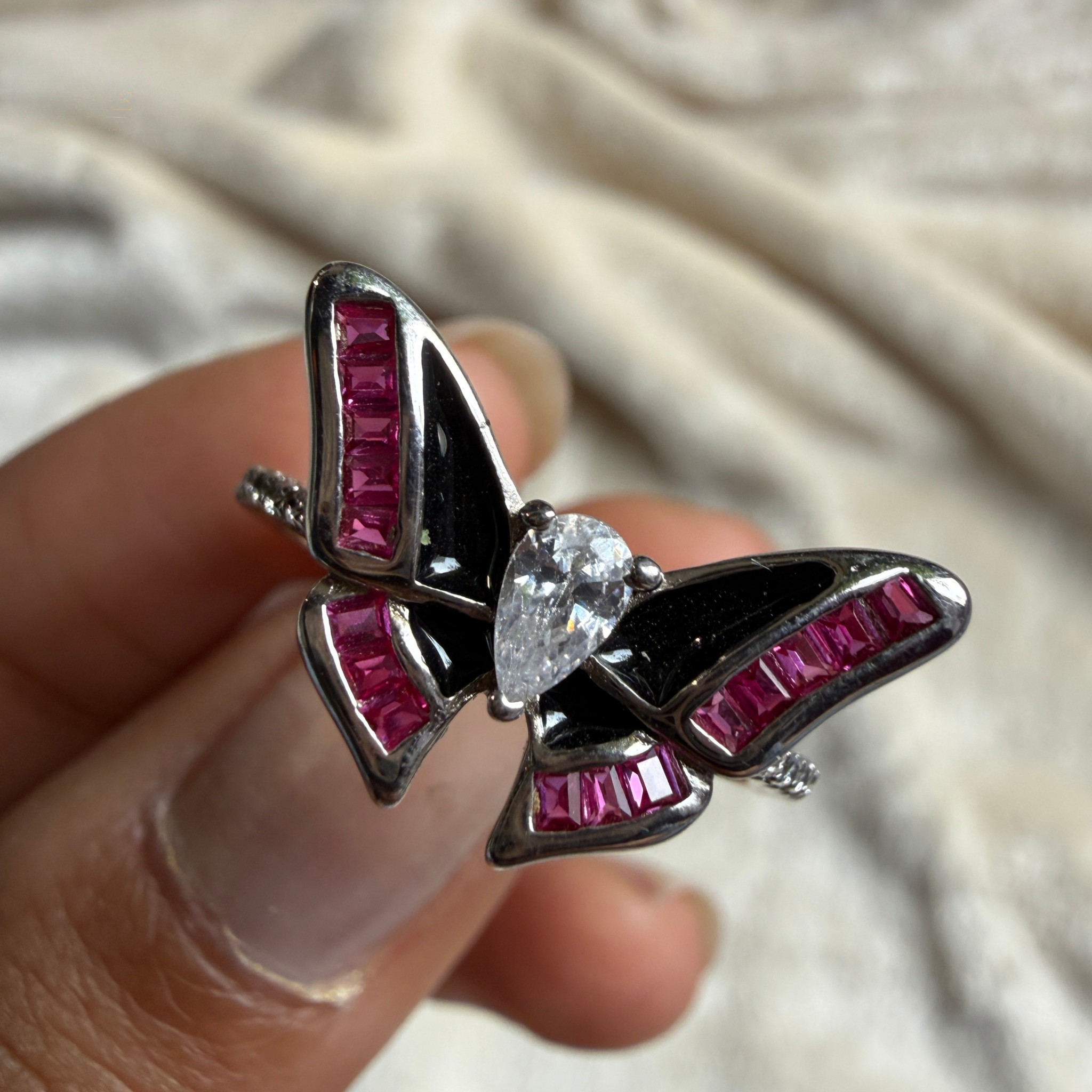 A close-up of a butterfly-shaped ring held between fingers. The ring features black enamel, silver-toned metal borders, and rows of bright pink baguette stones, with a clear teardrop-cut center stone.