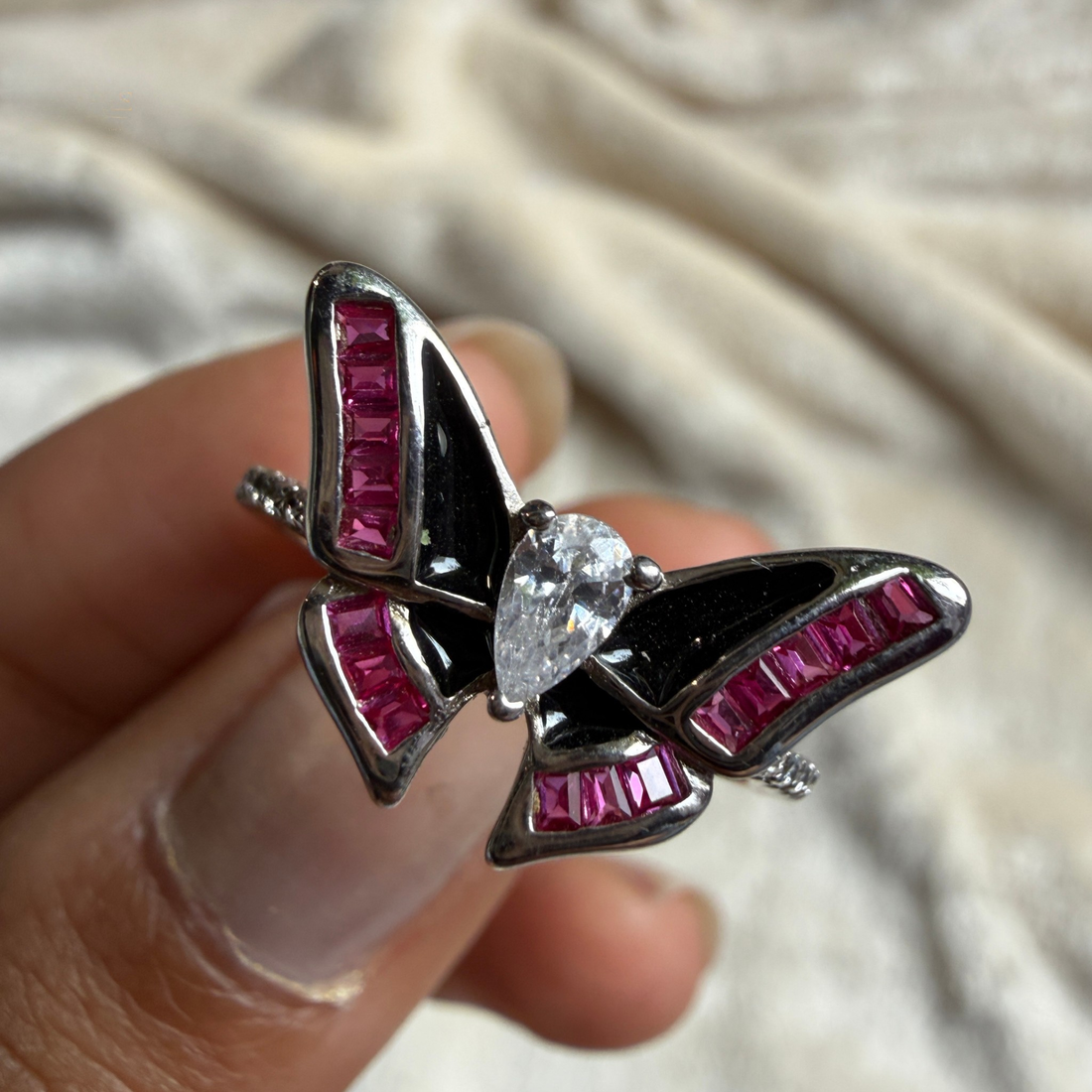 A close-up of a butterfly-shaped ring held between fingers. The ring features black enamel, silver-toned metal borders, and rows of bright pink baguette stones, with a clear teardrop-cut center stone.