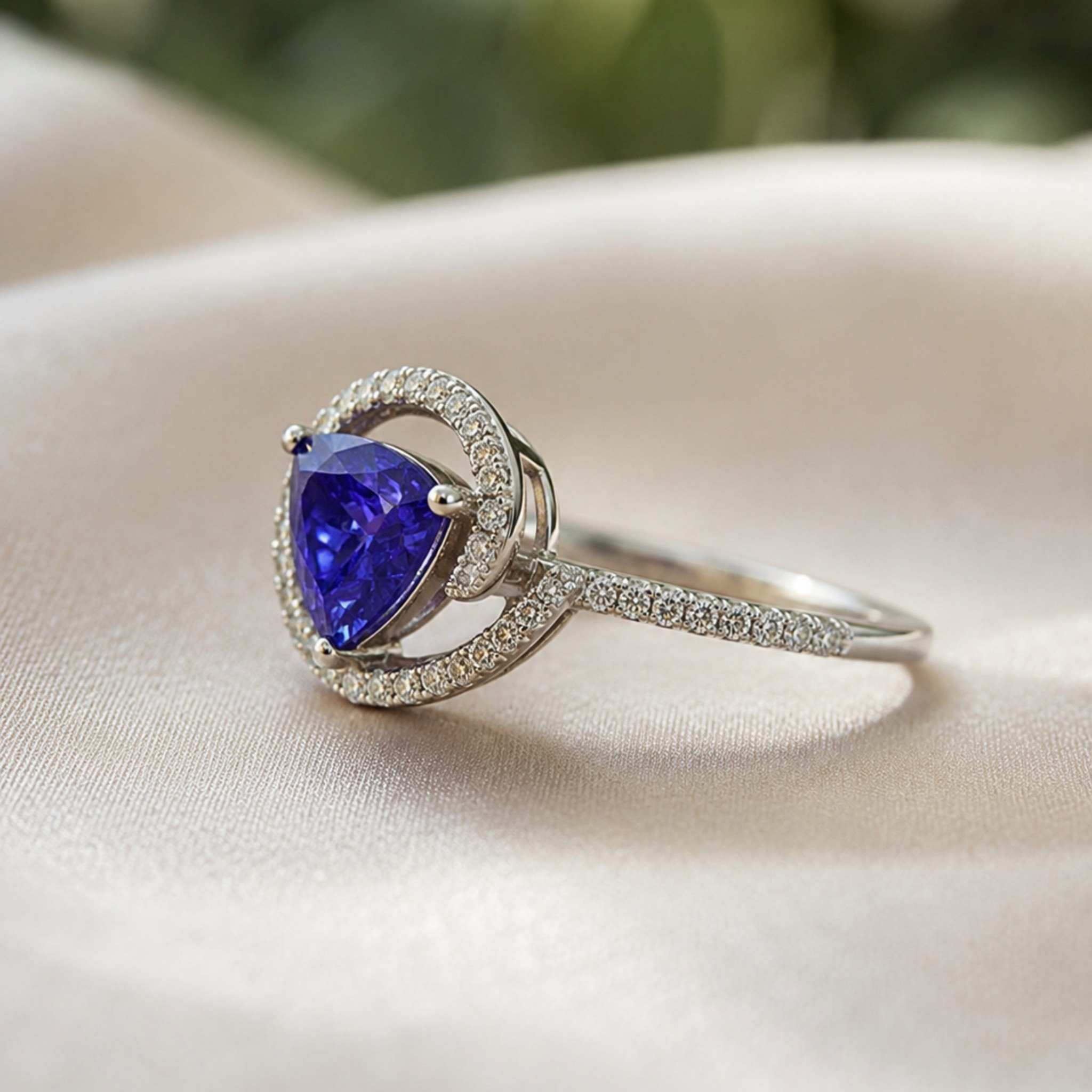 A silver ring featuring a pear-cut blue sapphire center stone and a halo of small clear stones, resting on a soft beige fabric surface.