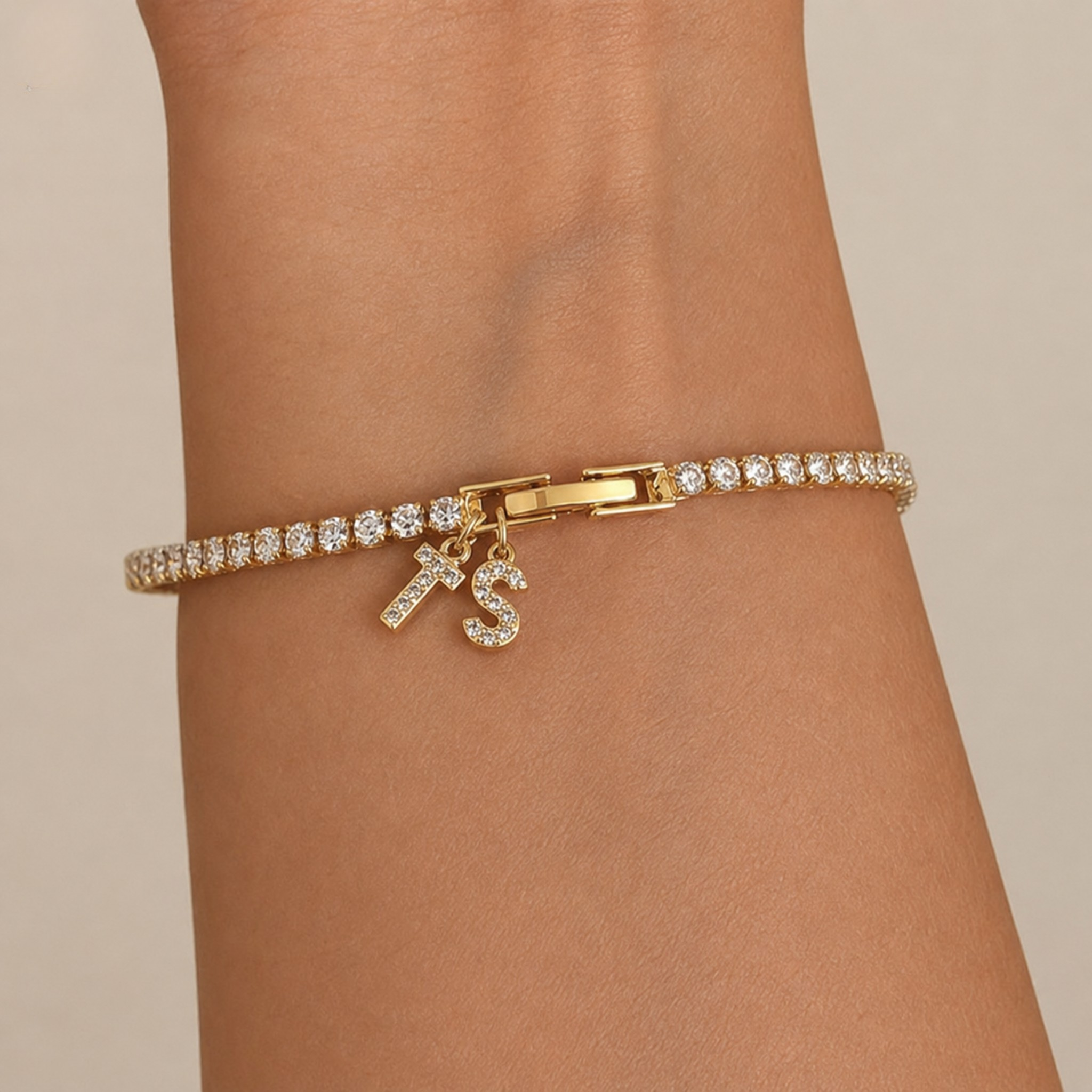 A gold bracelet with a row of clear stones and two dangling initial charms spelling 'KS', shown worn on a person's wrist against a plain beige background.