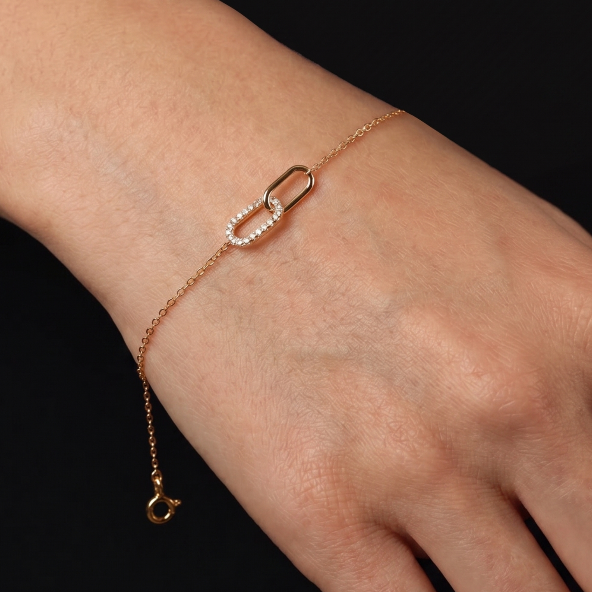 A delicate rose gold chain bracelet with a double interlocking link design, one of which is paved with small clear stones, shown worn on a hand against a solid black background.