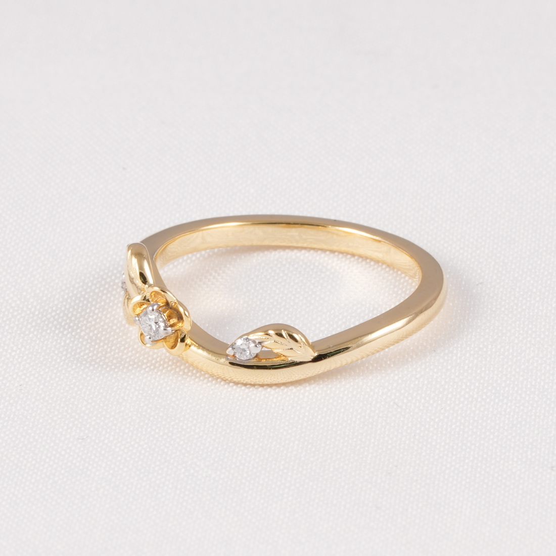 A gold ring featuring a small central clear gemstone and leaf-shaped details on either side, photographed on a textured white background.