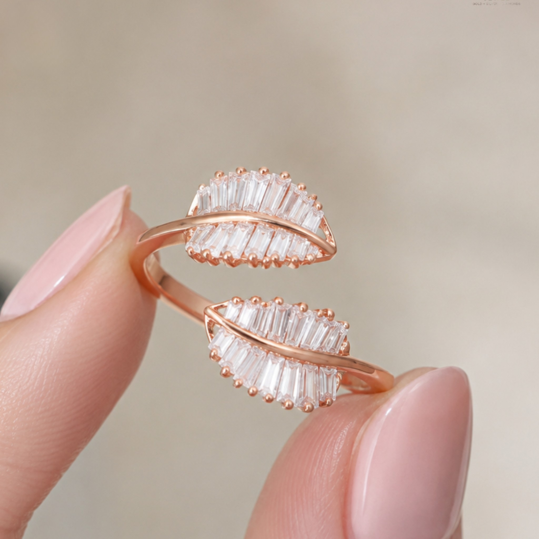 A close-up of a rose gold ring designed to look like two overlapping leaves, each adorned with clear baguette-cut crystals, held between fingers against a soft beige background.