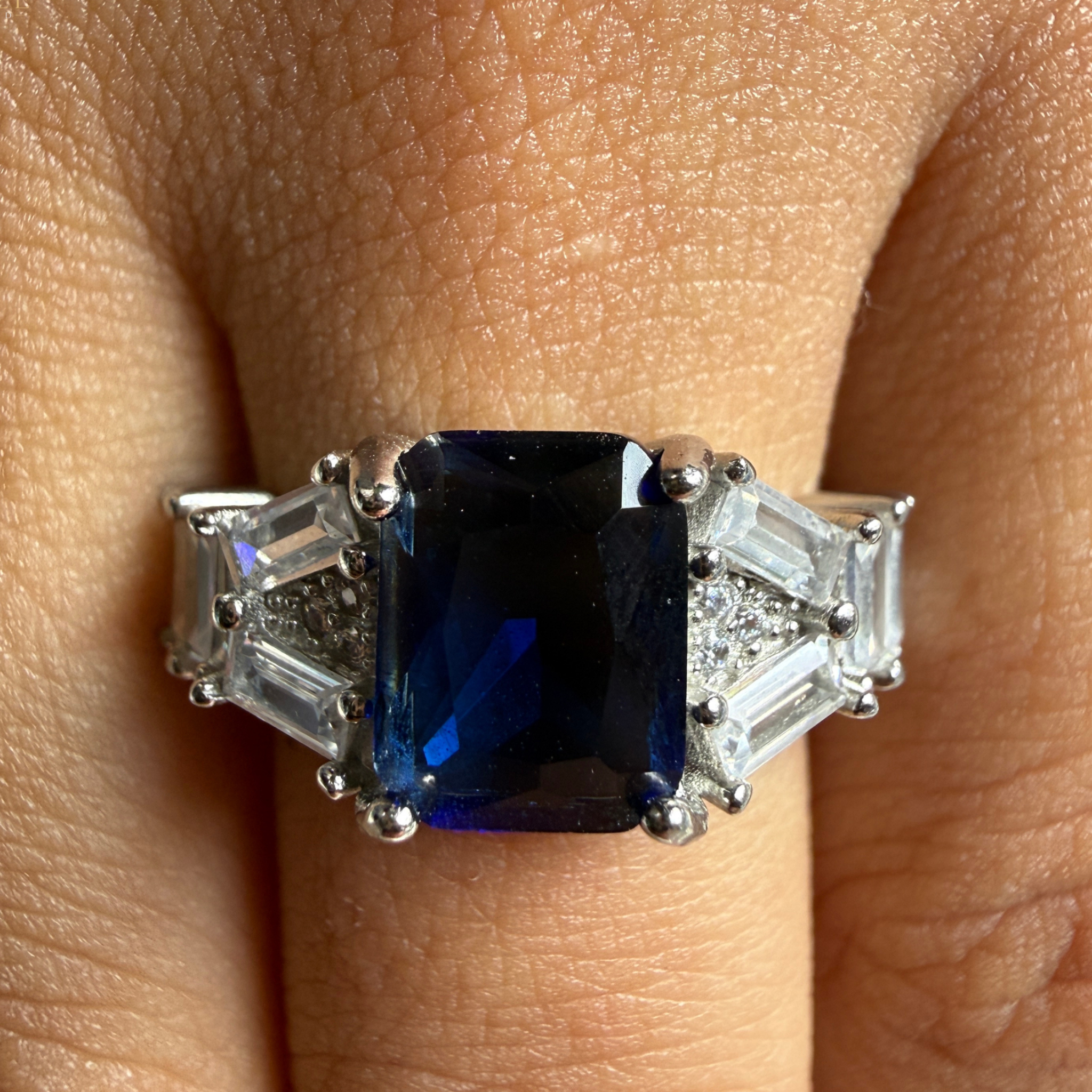 A silver ring with a large rectangular dark blue sapphire center stone and baguette side stones, shown worn on a finger.