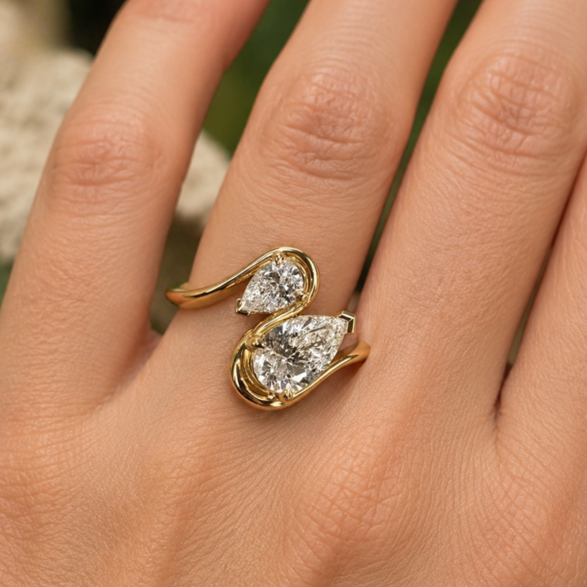 A close-up of a hand wearing a gold ring with two pear-cut clear gemstones in a curved, intertwined design.