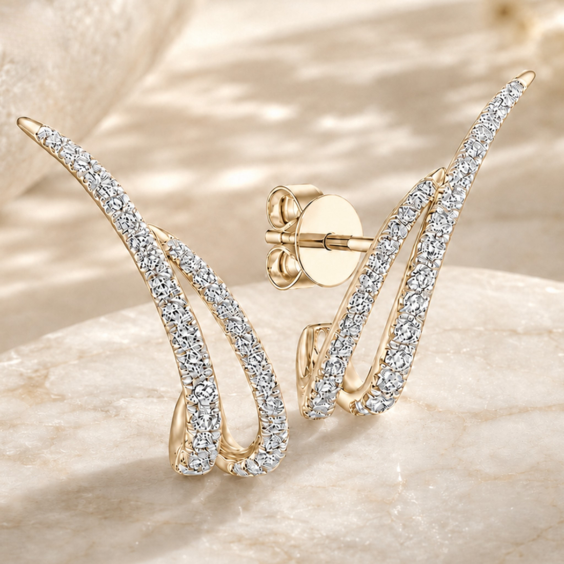 A pair of gold-plated earrings with curved, wing-like shapes paved with clear cubic zirconia stones, displayed on a beige marble surface with a gold post visible on one earring.