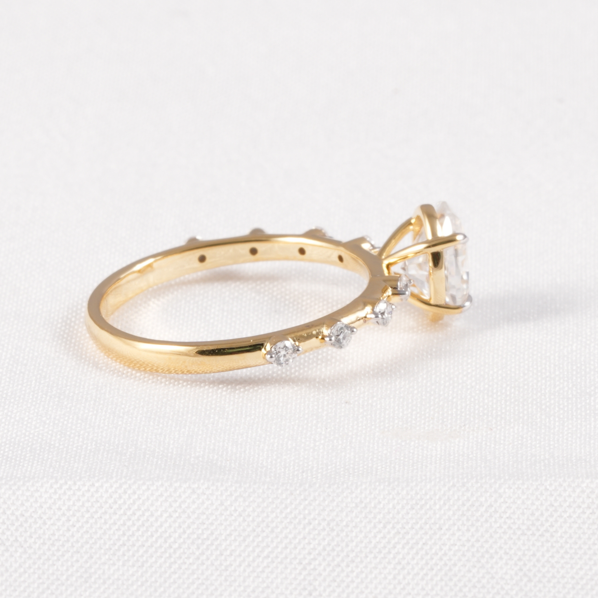 A yellow gold ring with a large clear gemstone and smaller clear stones along the band, photographed on a white textured fabric background.