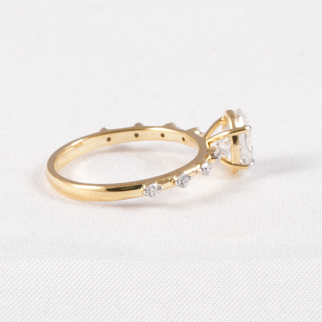 A yellow gold ring with a large clear gemstone and smaller clear stones along the band, photographed on a white textured fabric background.