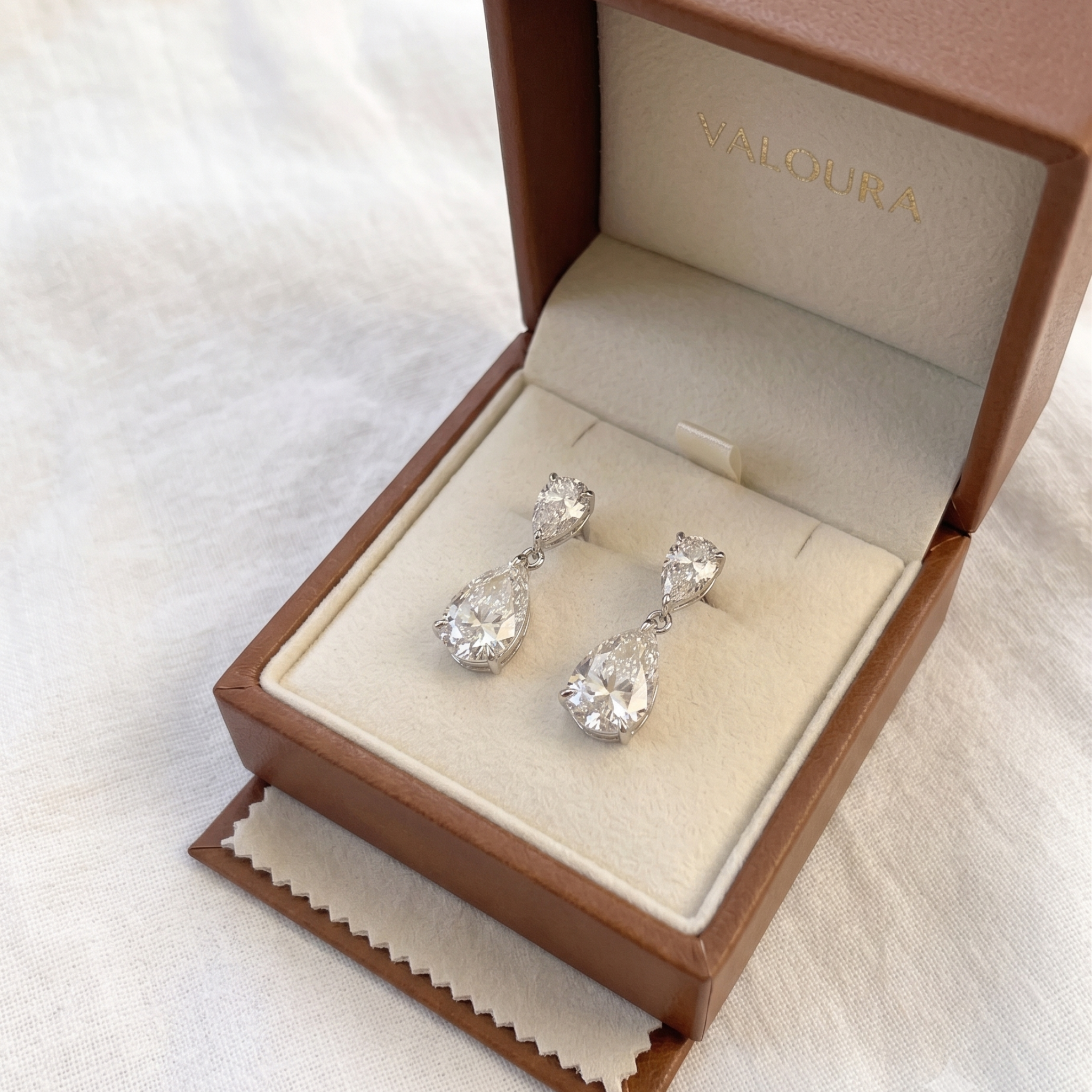 A pair of silver teardrop dangle earrings with clear gemstones, presented in an open brown Saloura jewelry box on a white fabric background.