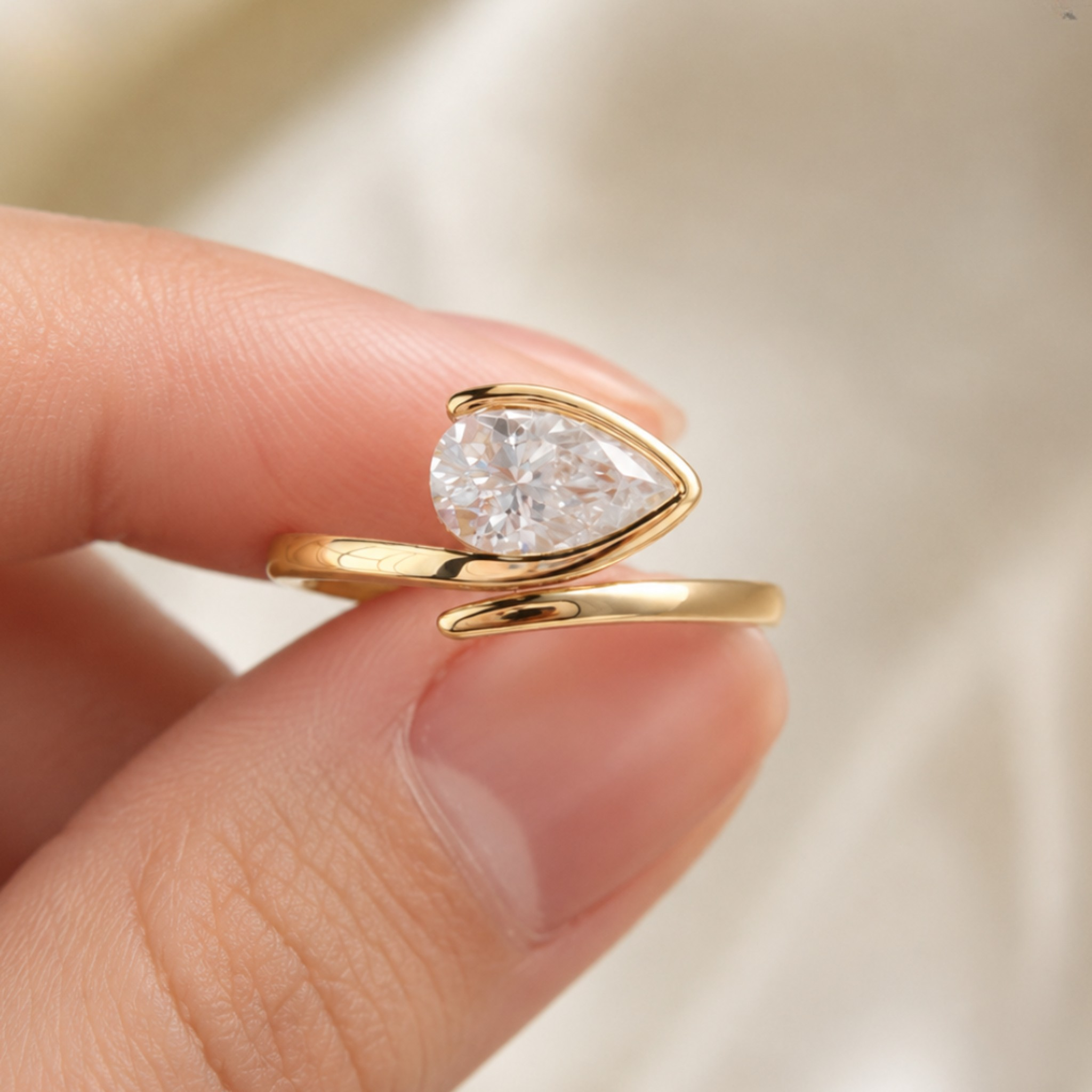A gold ring with a pear-cut clear gemstone held between two fingers against a soft, light beige background.