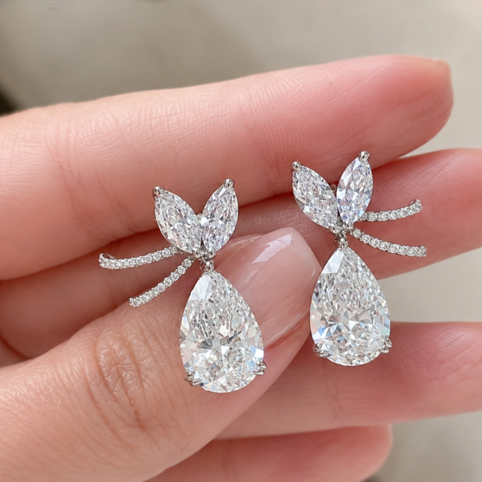 A pair of pear-cut diamond drop earrings with marquise-cut diamond bow details and small round diamonds on the posts, held between fingers to show scale.