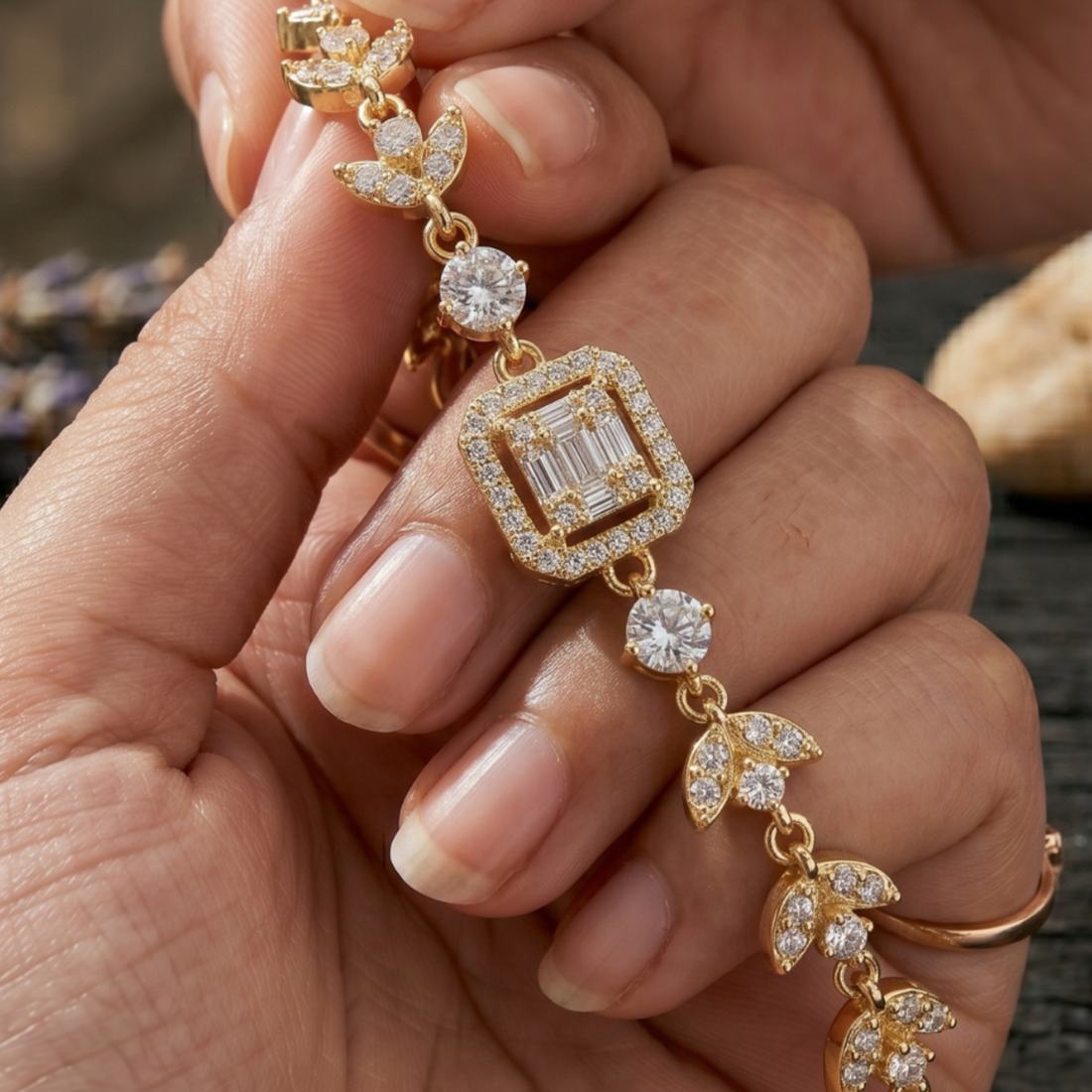 A close-up of a hand holding a gold bracelet with a central square baguette diamond surrounded by small diamonds, connected by leaf-shaped and round diamond details.