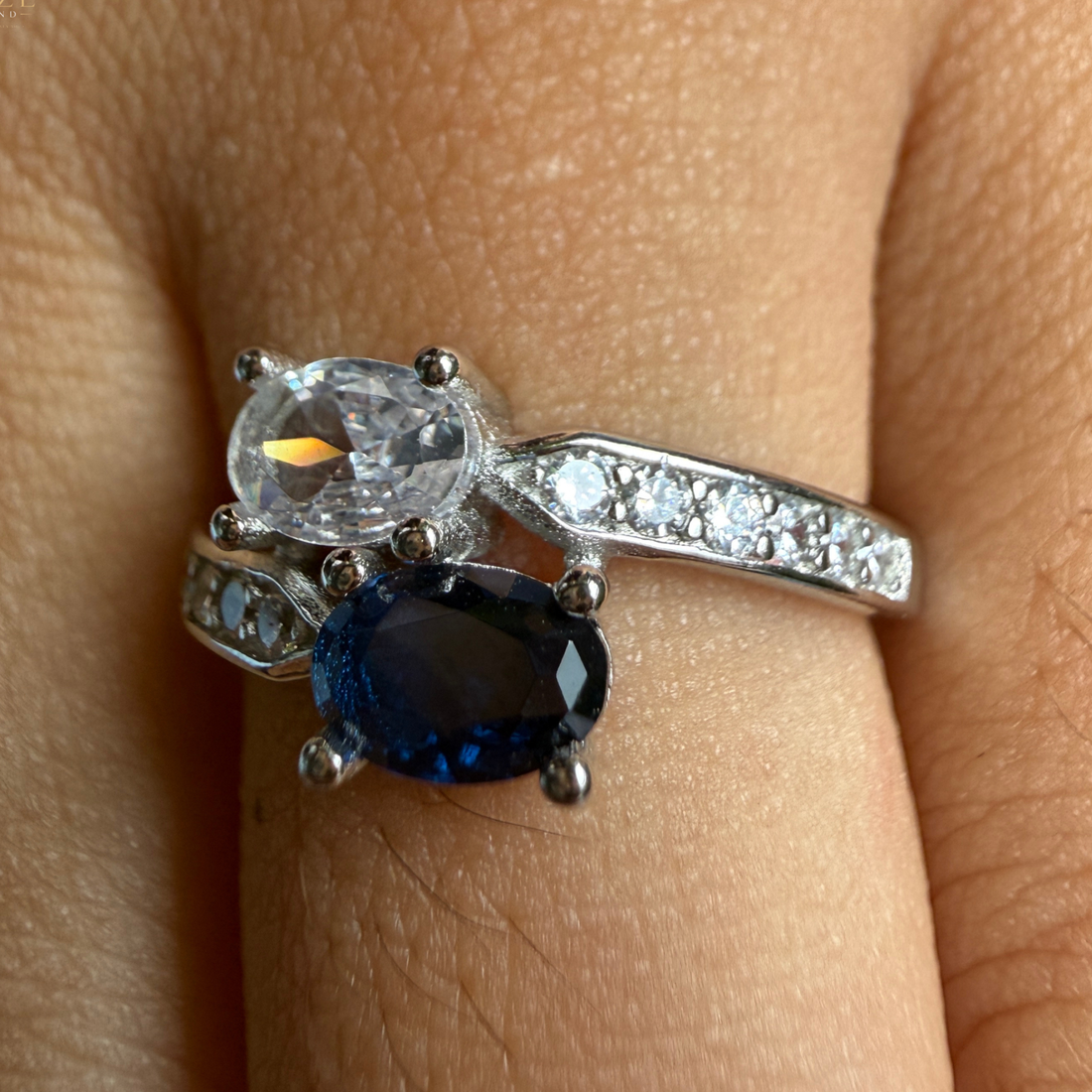 A close-up of a ring worn on a finger, featuring a silver-toned band with clear pavé stones and two oval gemstones: a large clear marquise-cut stone on top and a larger deep blue oval stone below.