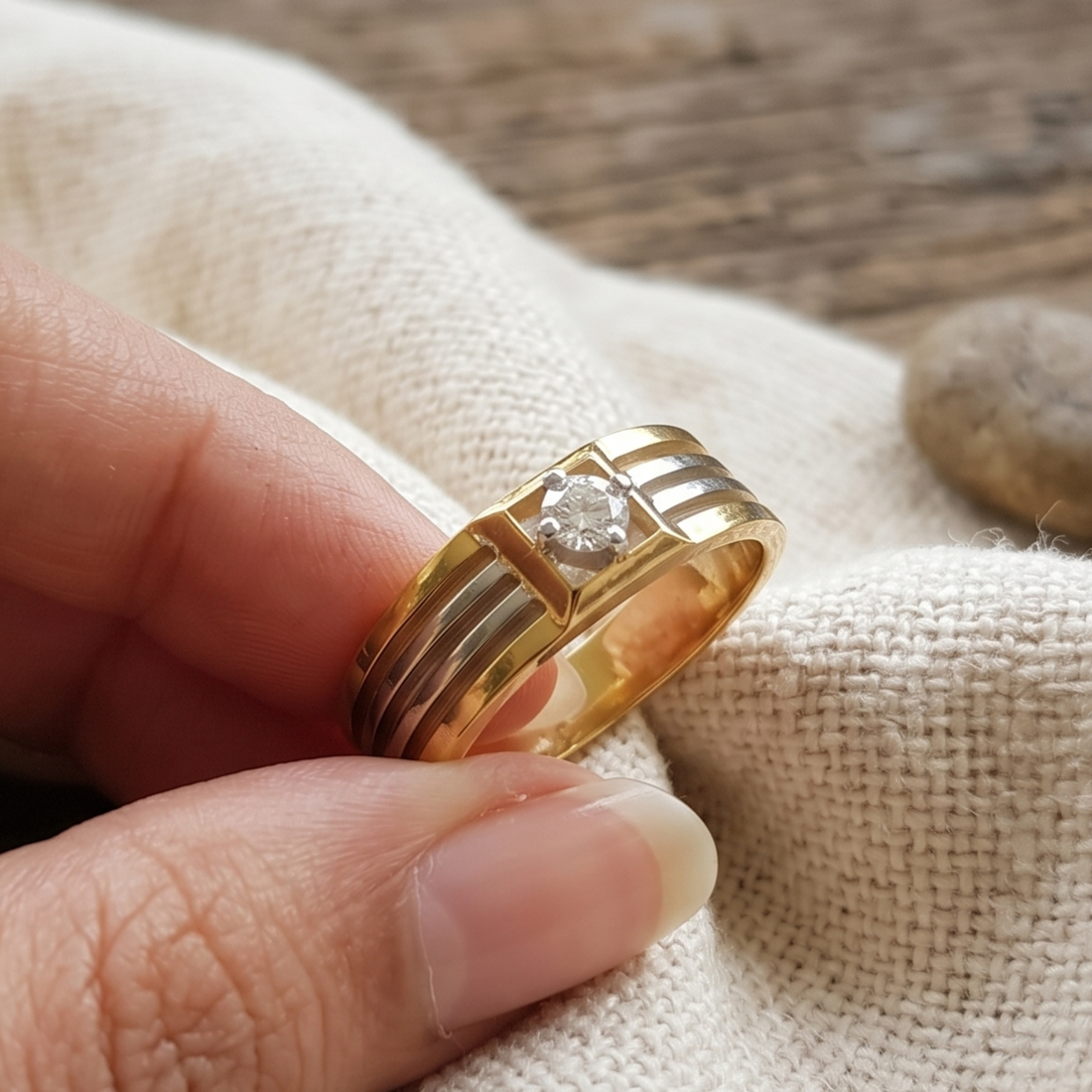 A gold ring with a single round clear gemstone held between fingers over a textured beige fabric background.