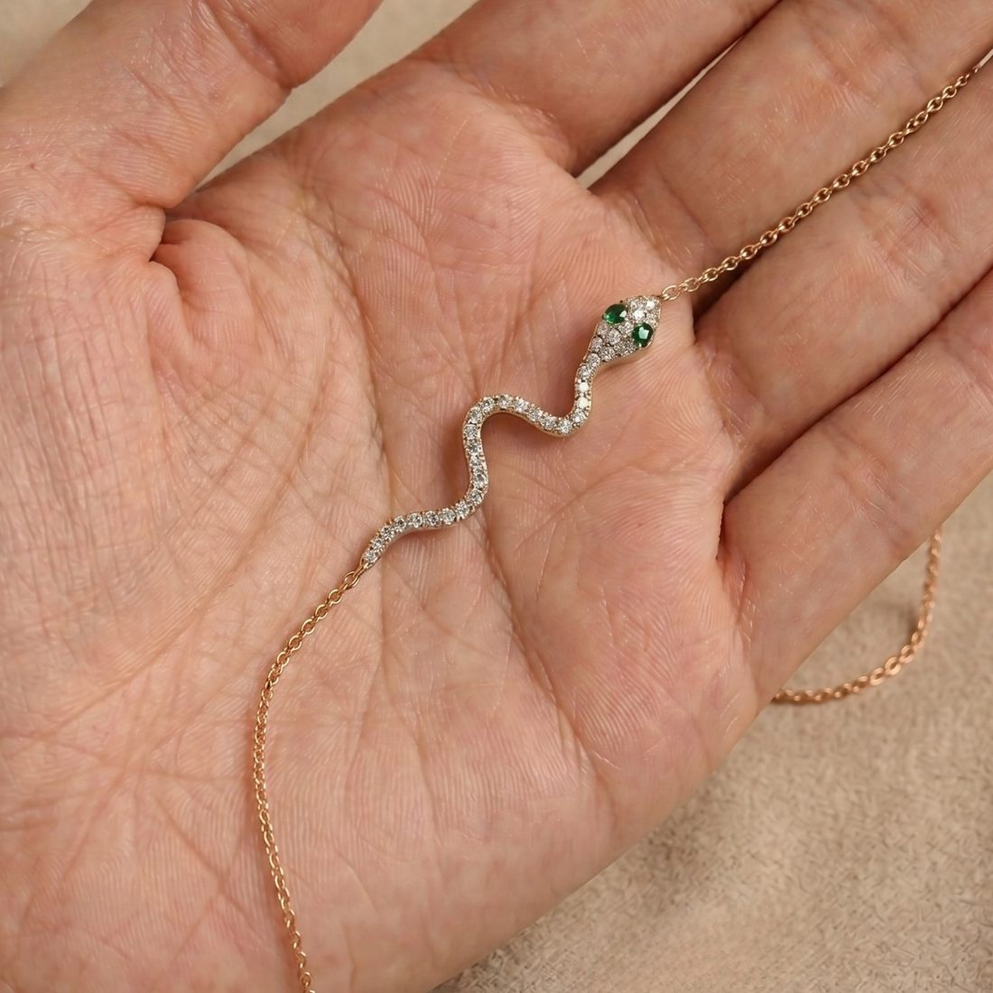 A delicate snake necklace with a rose gold chain, clear pavé-set stones, and a green gemstone head, displayed on a person's palm.