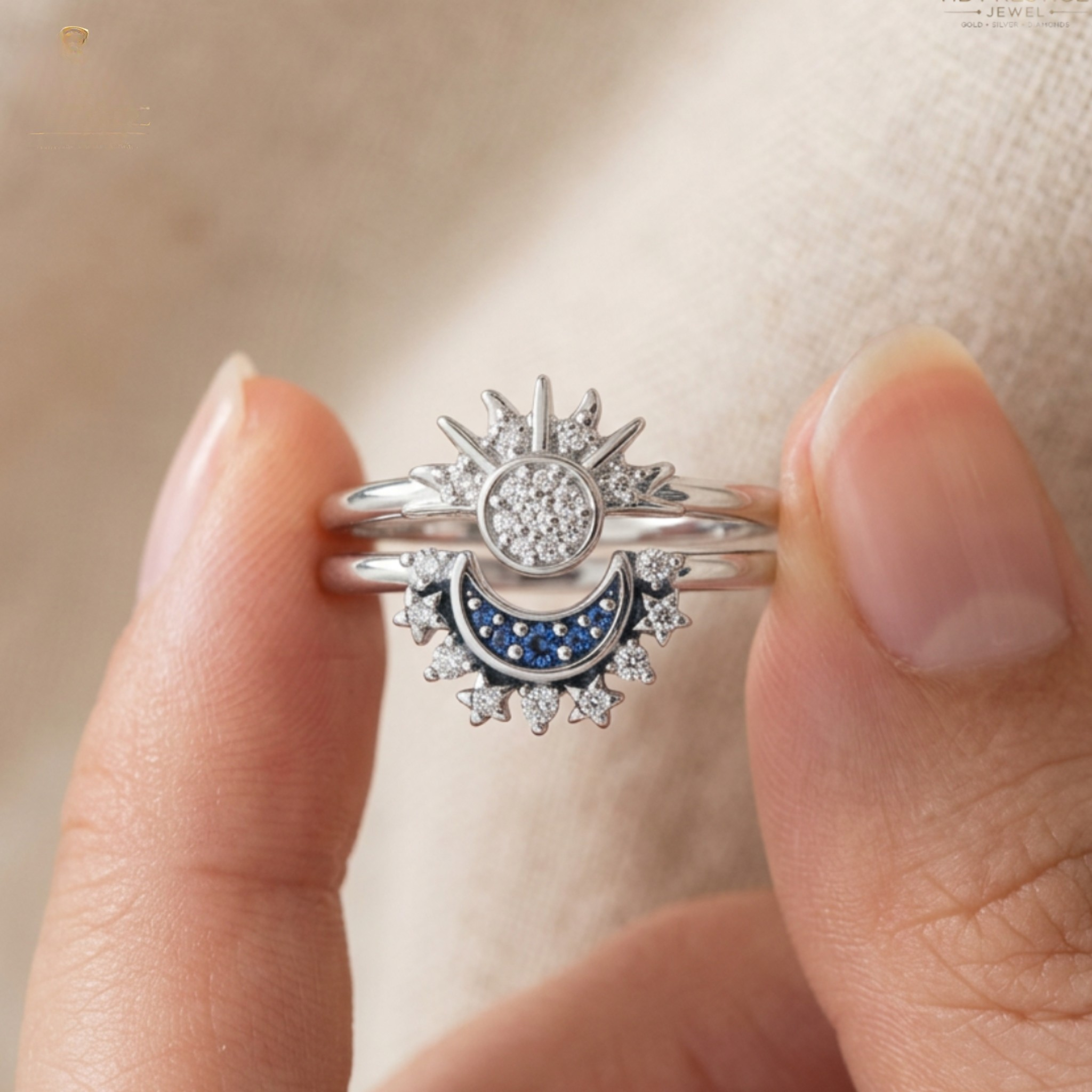 A close-up of a hand holding a silver-toned double ring set featuring a sunburst and crescent moon design with clear stones and a blue crescent inlay.