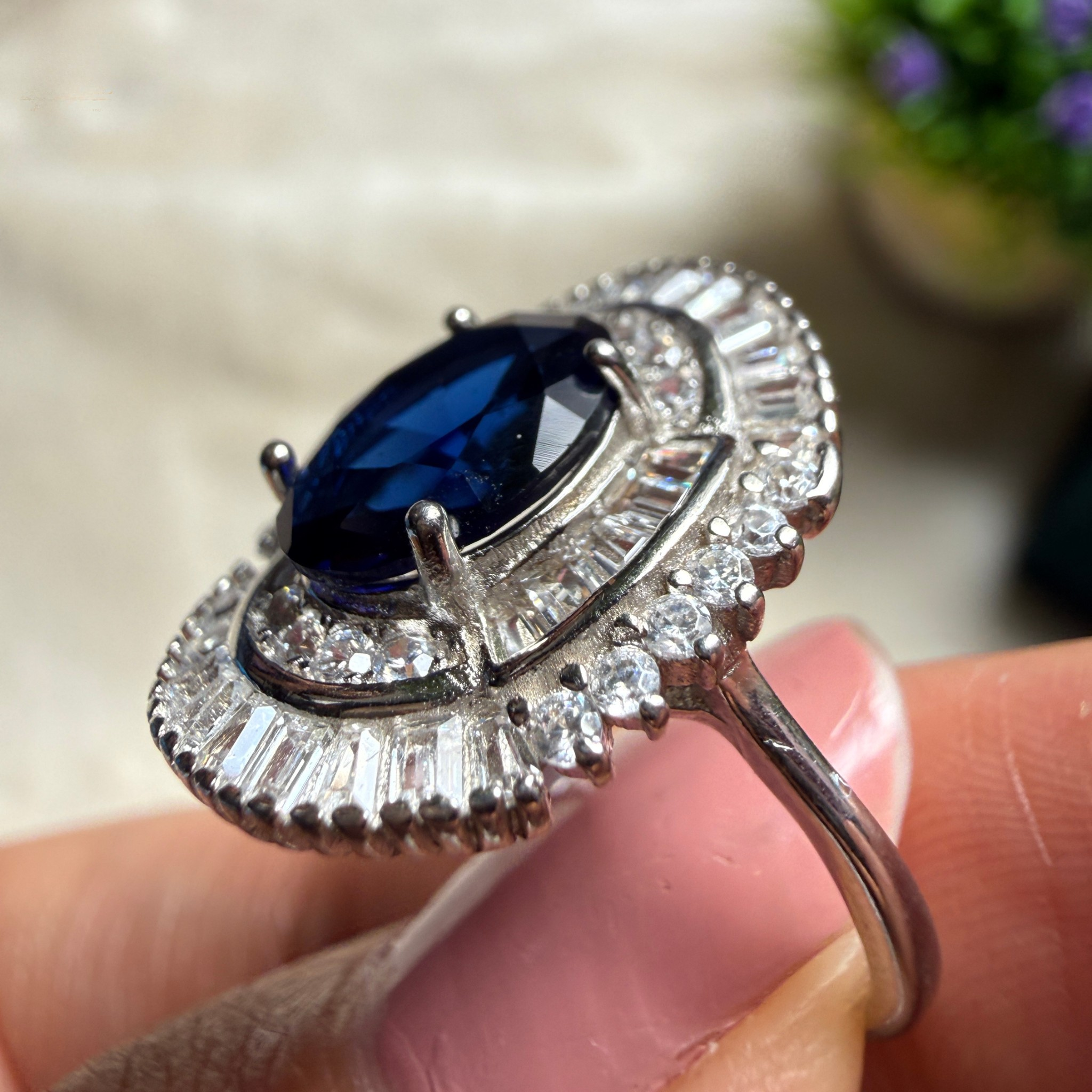A silver-toned halo ring featuring a large oval deep blue sapphire center stone and surrounding baguette-cut clear stones, held between fingers against a softly blurred light background.