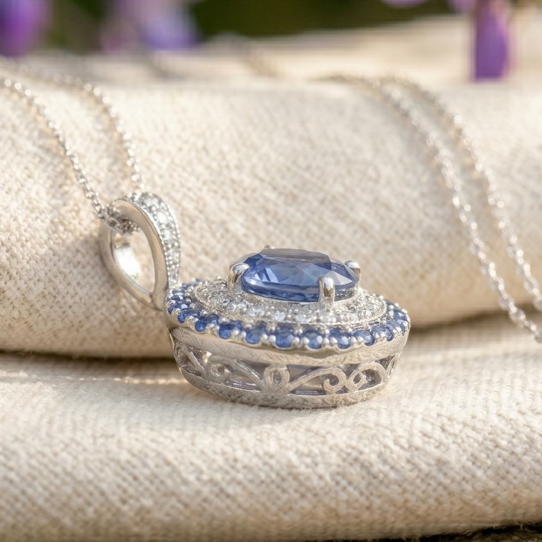 A silver-toned oval pendant necklace with a central blue gemstone and small blue accent stones, displayed on a textured beige fabric background.