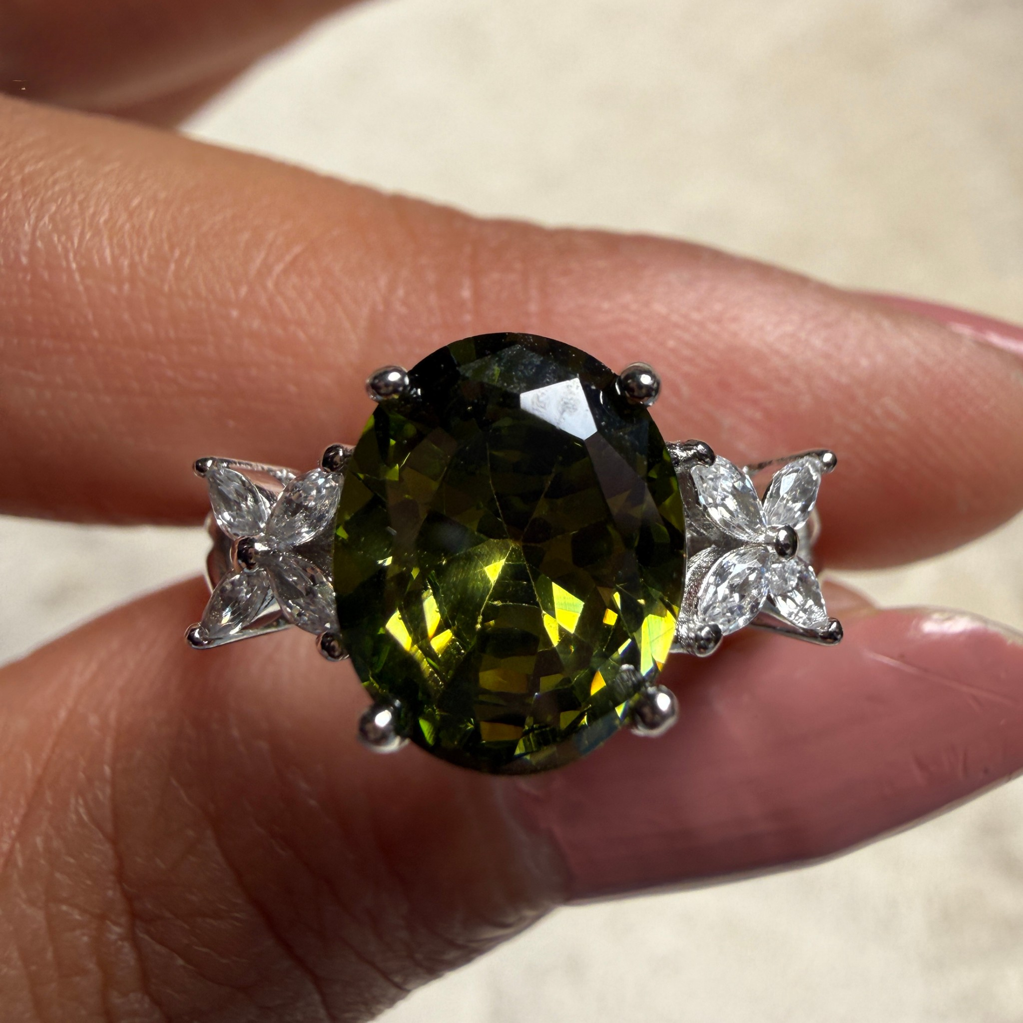 A close-up of a ring being worn on a person's finger, featuring a large oval faceted olive-green gemstone in a silver-toned setting with small clear accent stones on each side.