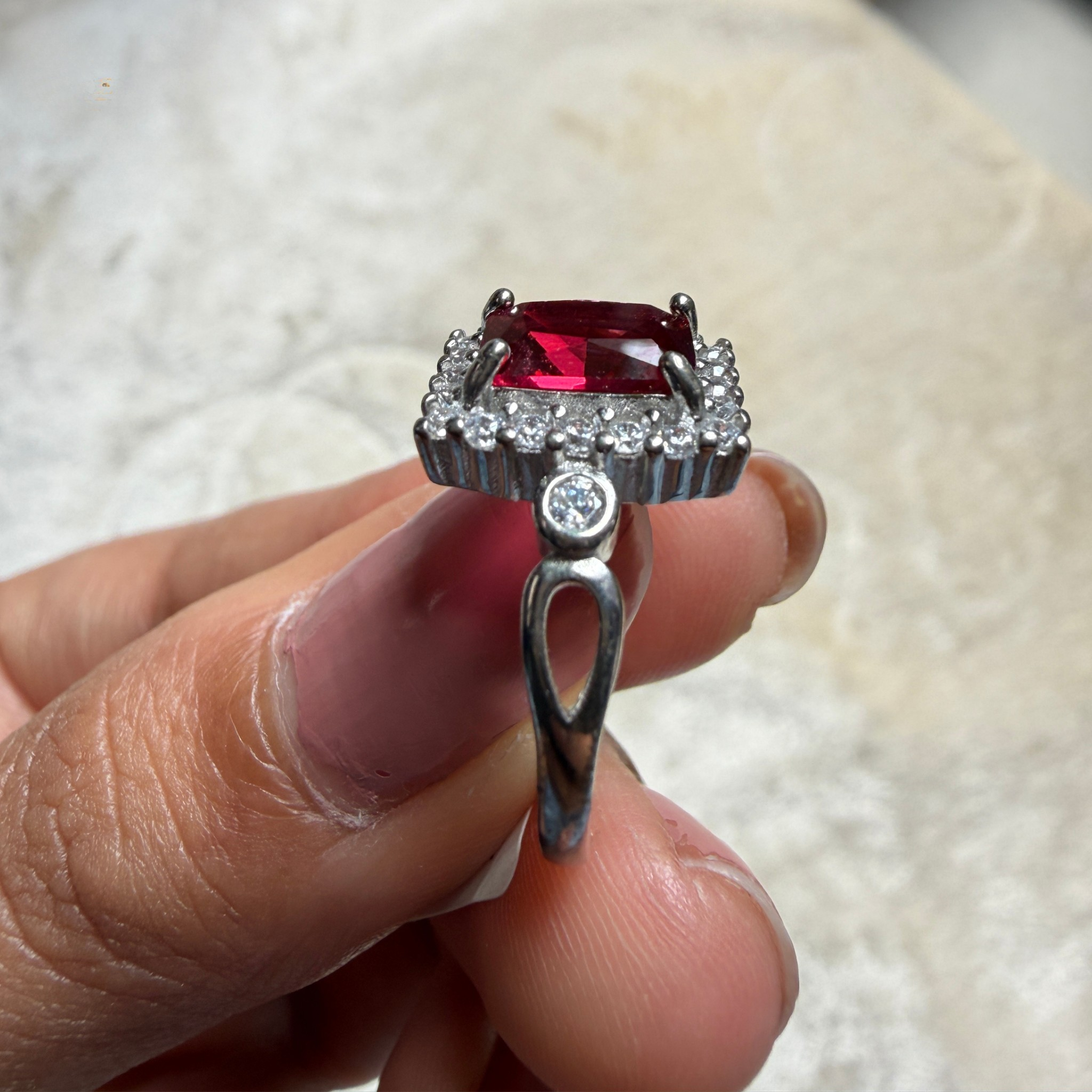 A silver ring with a rectangular red gemstone and a halo of small clear stones, held between fingers against a light textured background.