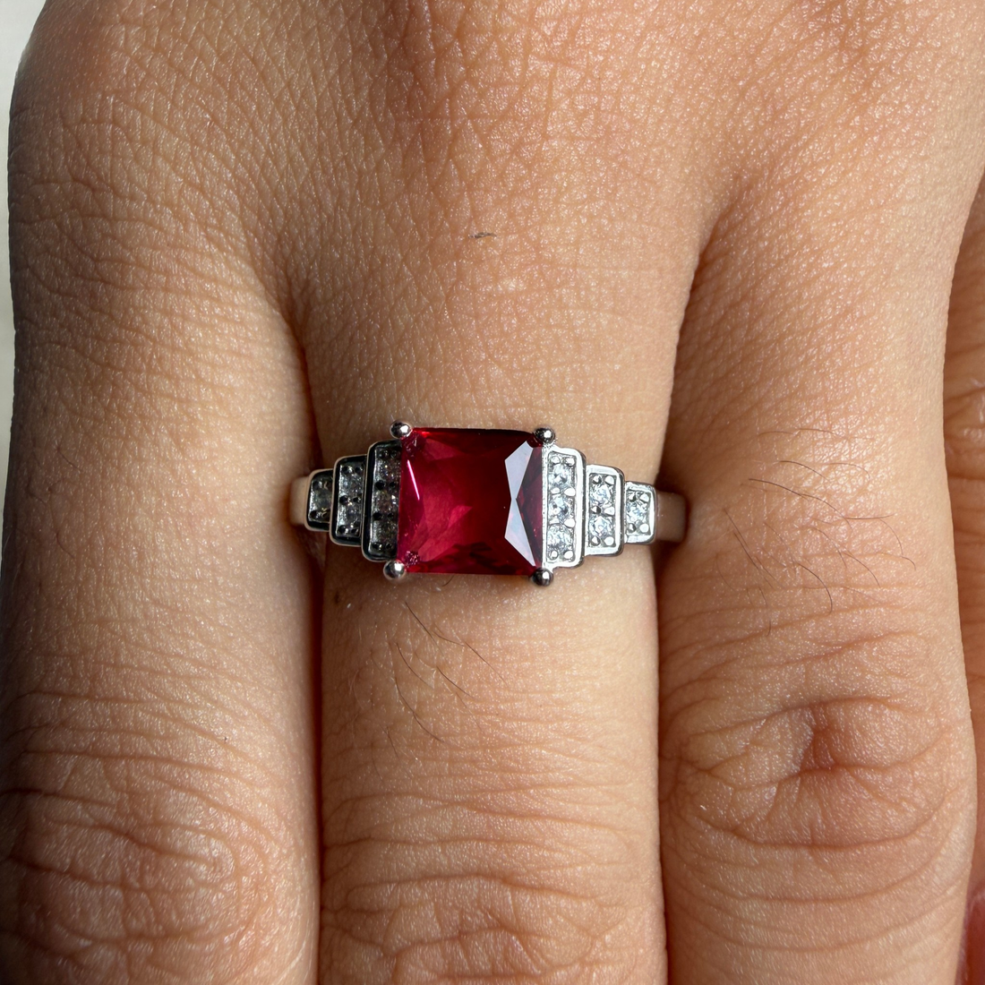 A silver ring with a rectangular red gemstone and small clear accent stones, shown worn on a finger.