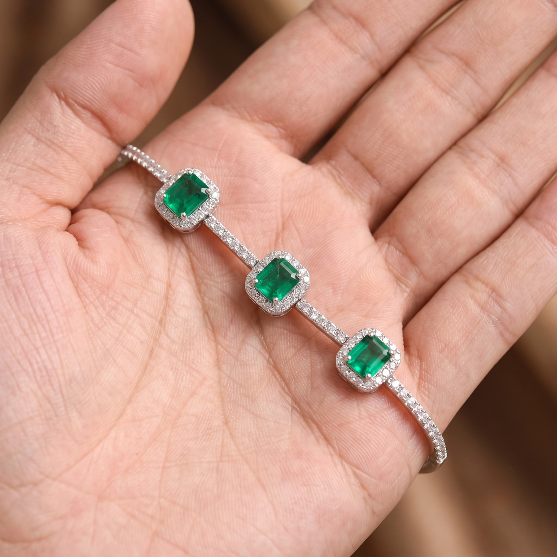 A silver-toned three-stone bracelet with square-cut green emerald-cut gemstones and halo settings, displayed on an open hand.