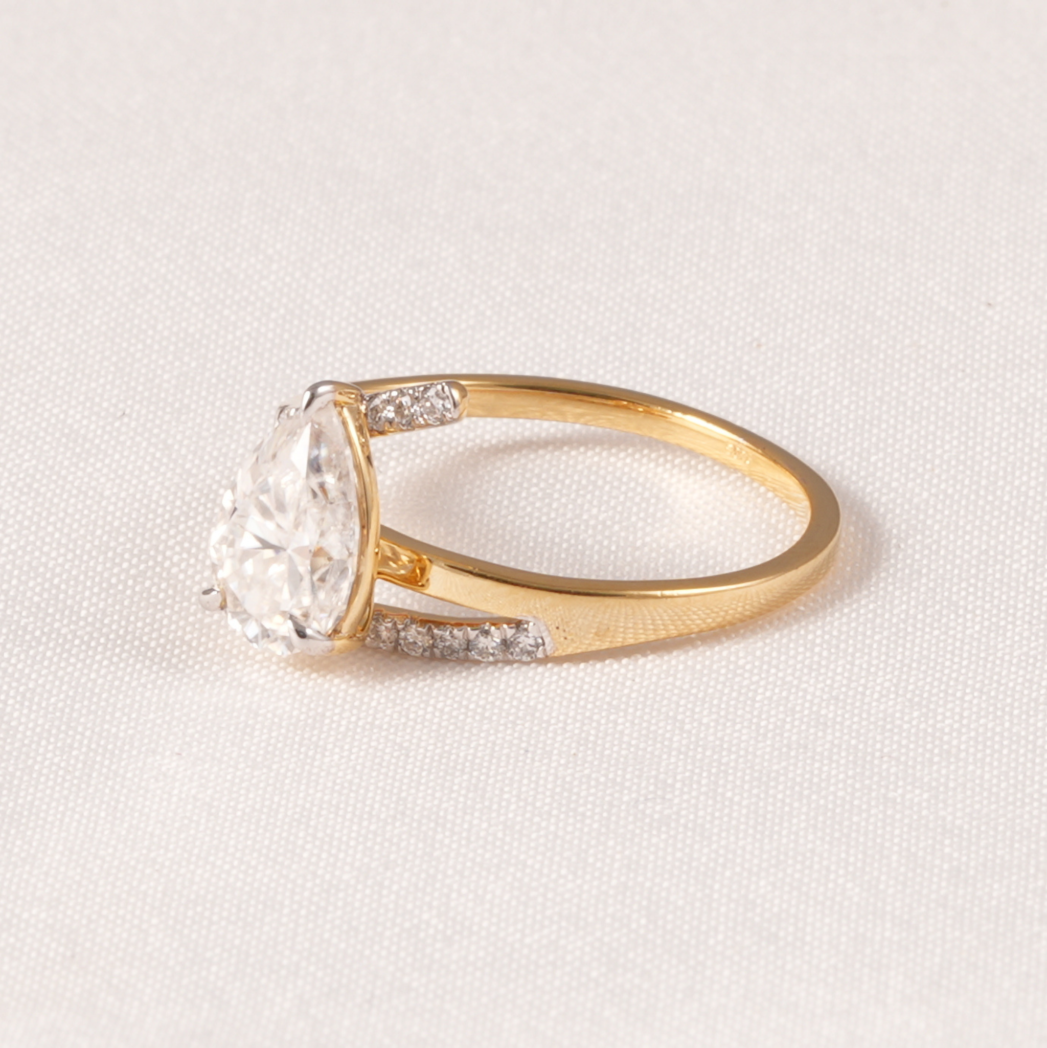 A yellow gold ring featuring a pear-cut clear center stone and small pavé-set accent stones on the band, photographed on a textured off-white fabric background.