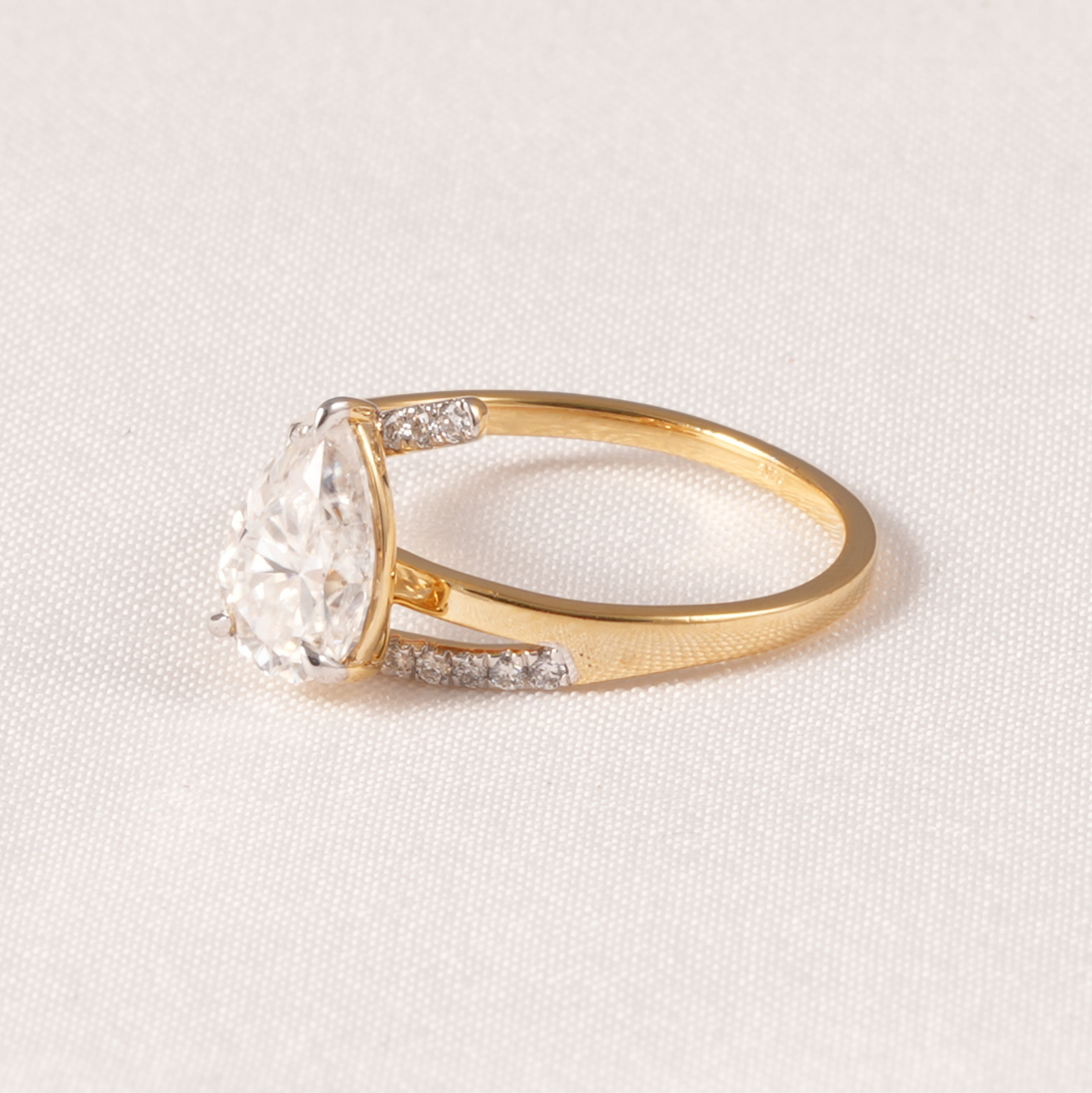 A yellow gold ring featuring a pear-cut clear center stone and small pavé-set accent stones on the band, photographed on a textured off-white fabric background.