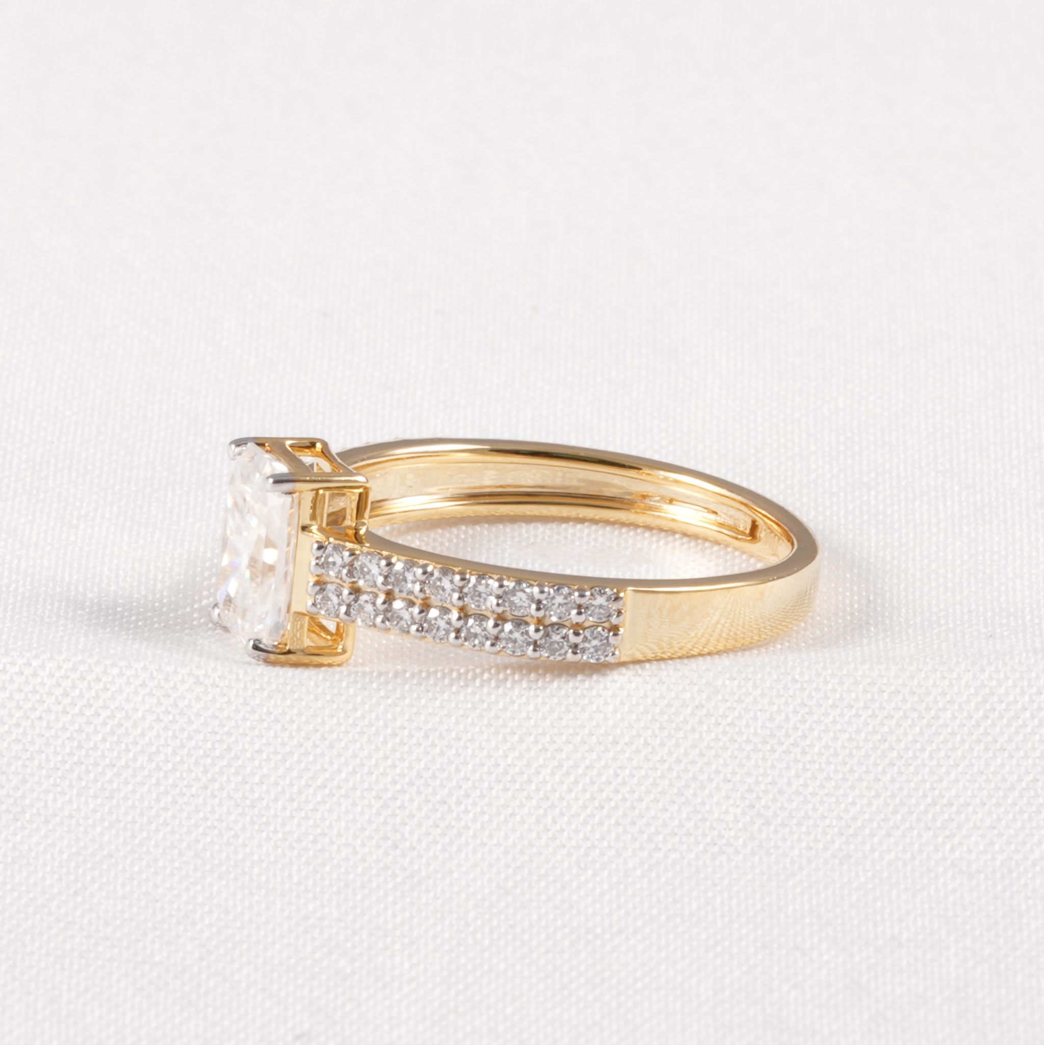 A yellow gold ring featuring a large rectangular clear gemstone in a prong setting, with a pavé band accented by small clear stones, photographed on a textured white fabric background.